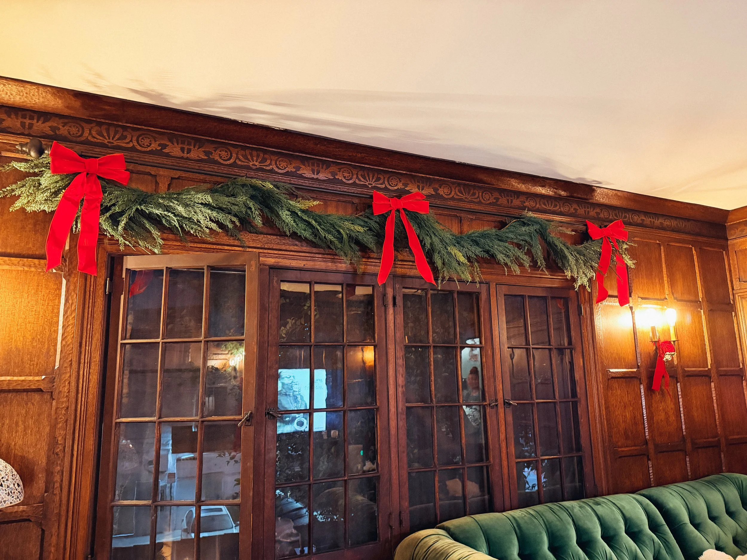Christmas garland with red bows hanging above wooden framed glass doors in a room with wood-paneled walls and a green velvet sofa.