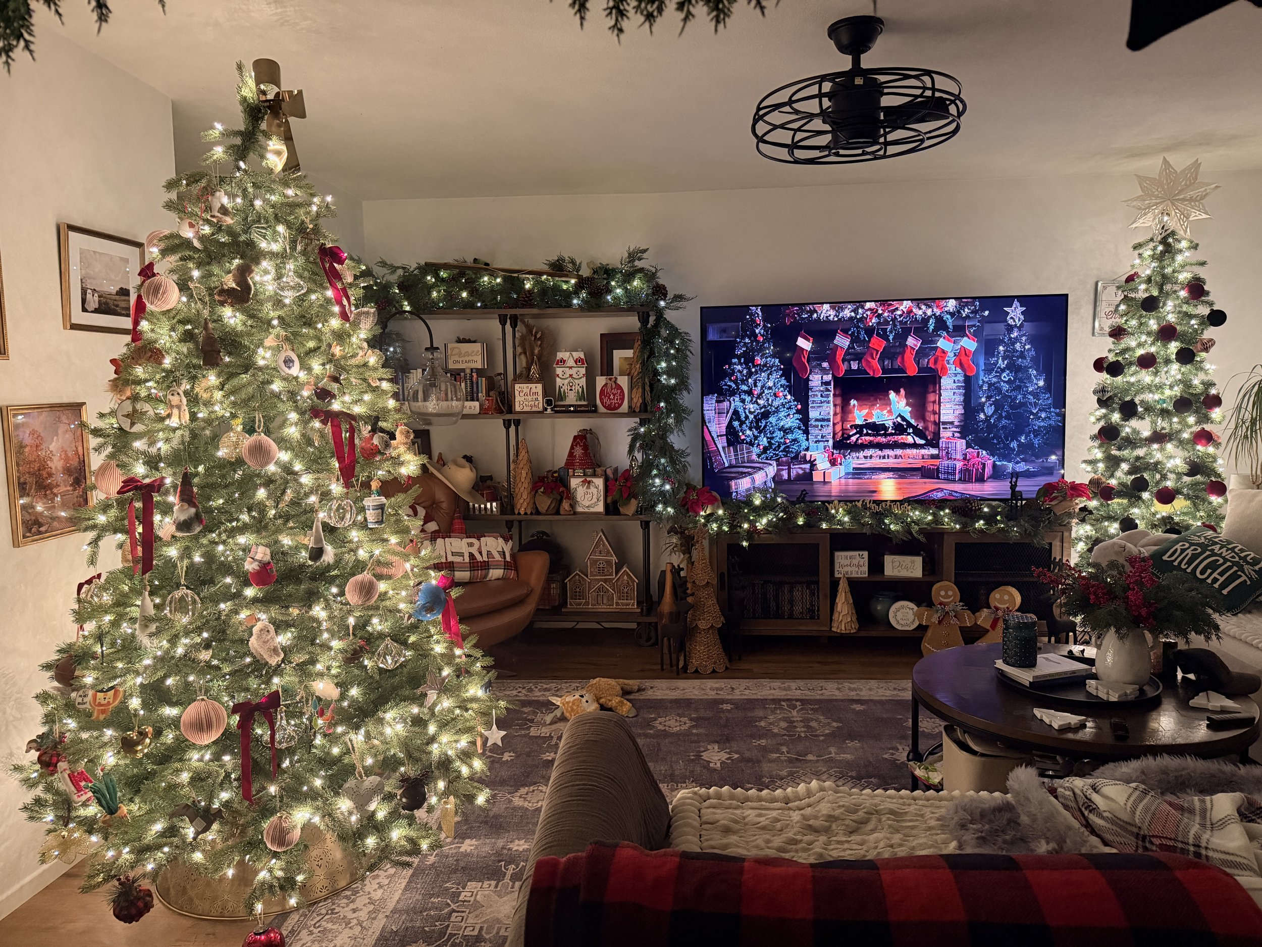 Living room decorated for Christmas with two decorated Christmas trees, a fireplace on TV screen, and holiday decorations.