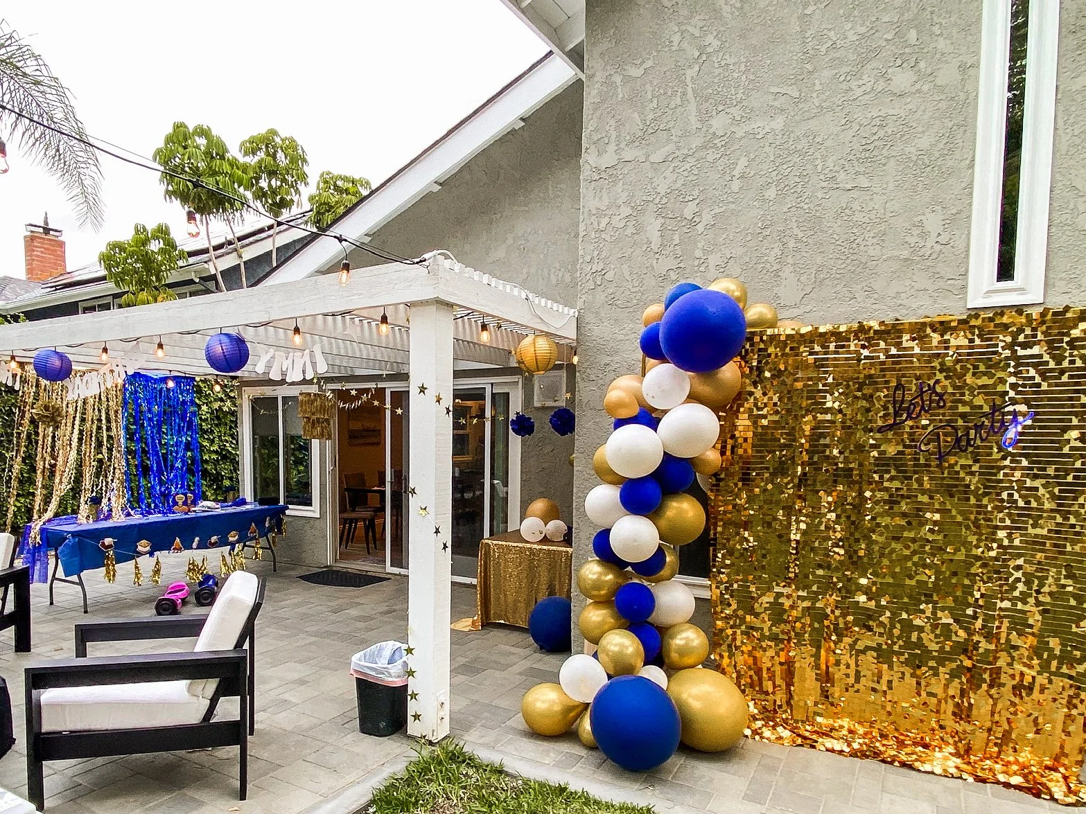 Decorations for a party in a backyard with blue, gold, and white balloons, a gold sequin backdrop with 