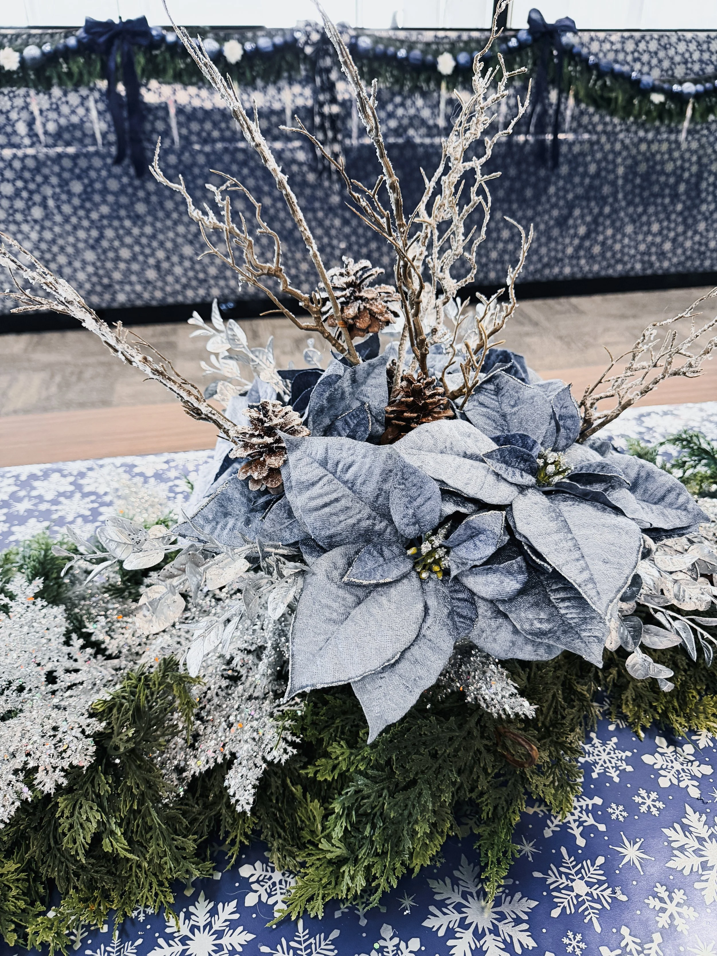 A winter-themed floral arrangement with silver leaves, pinecones, and frosted branches on a blue tablecloth with white snowflake patterns.