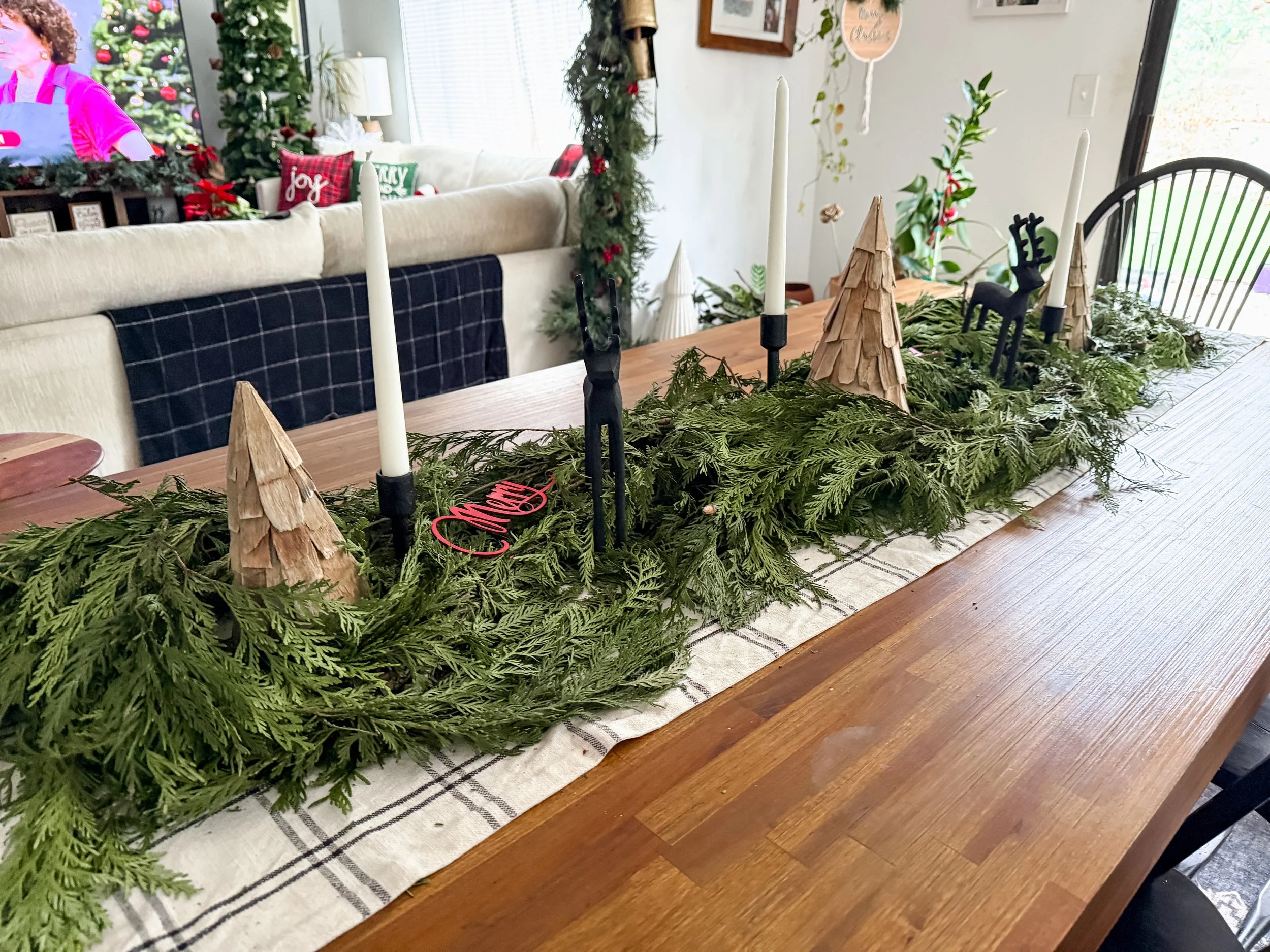 Christmas centerpiece on a wooden dining table with greenery, small wooden trees, black reindeer and candle holders.