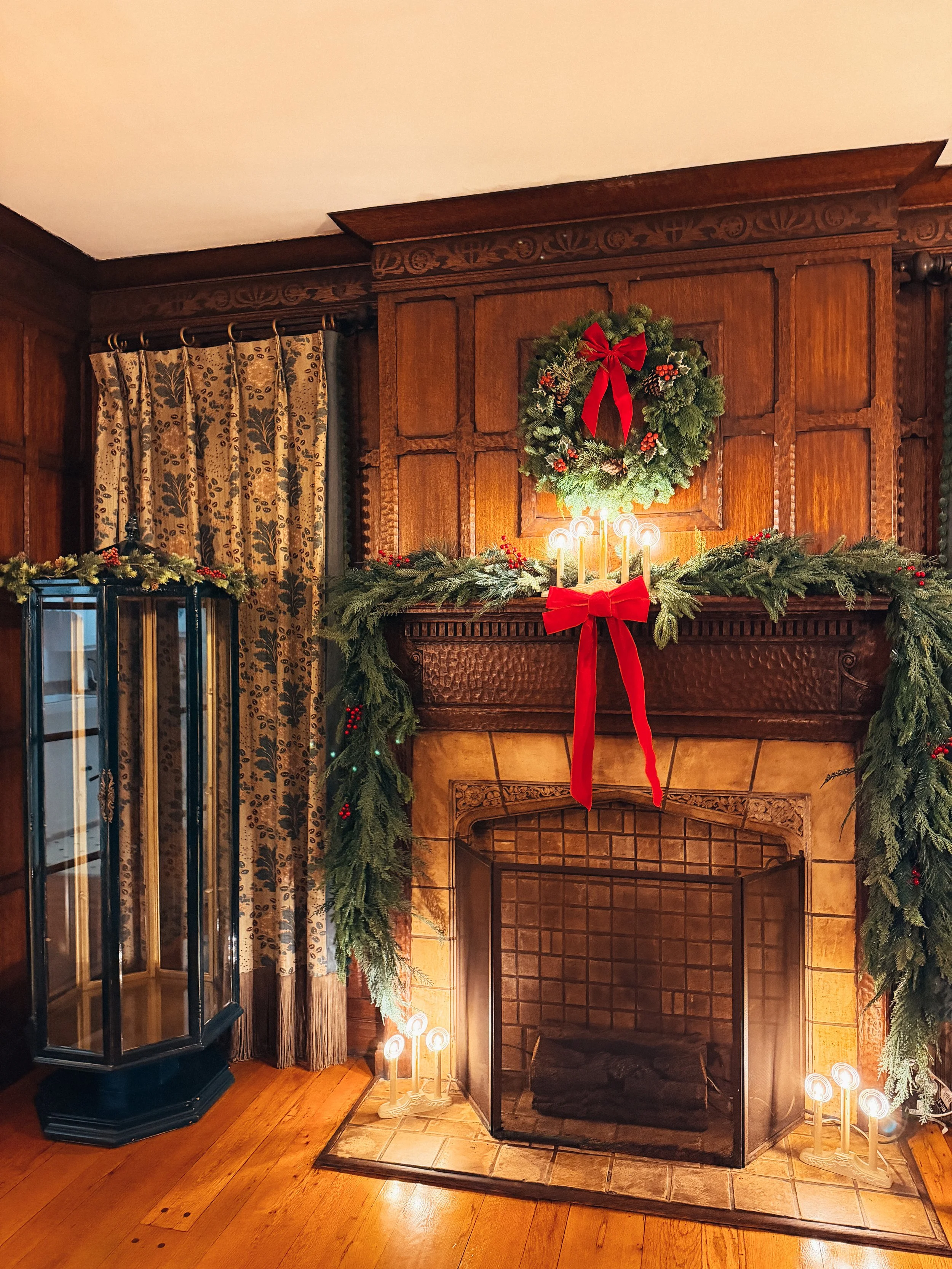 Decorated Christmas fireplace with a green wreath with a red bow, garland with red berries, and candles, in a cozy wood-paneled room.