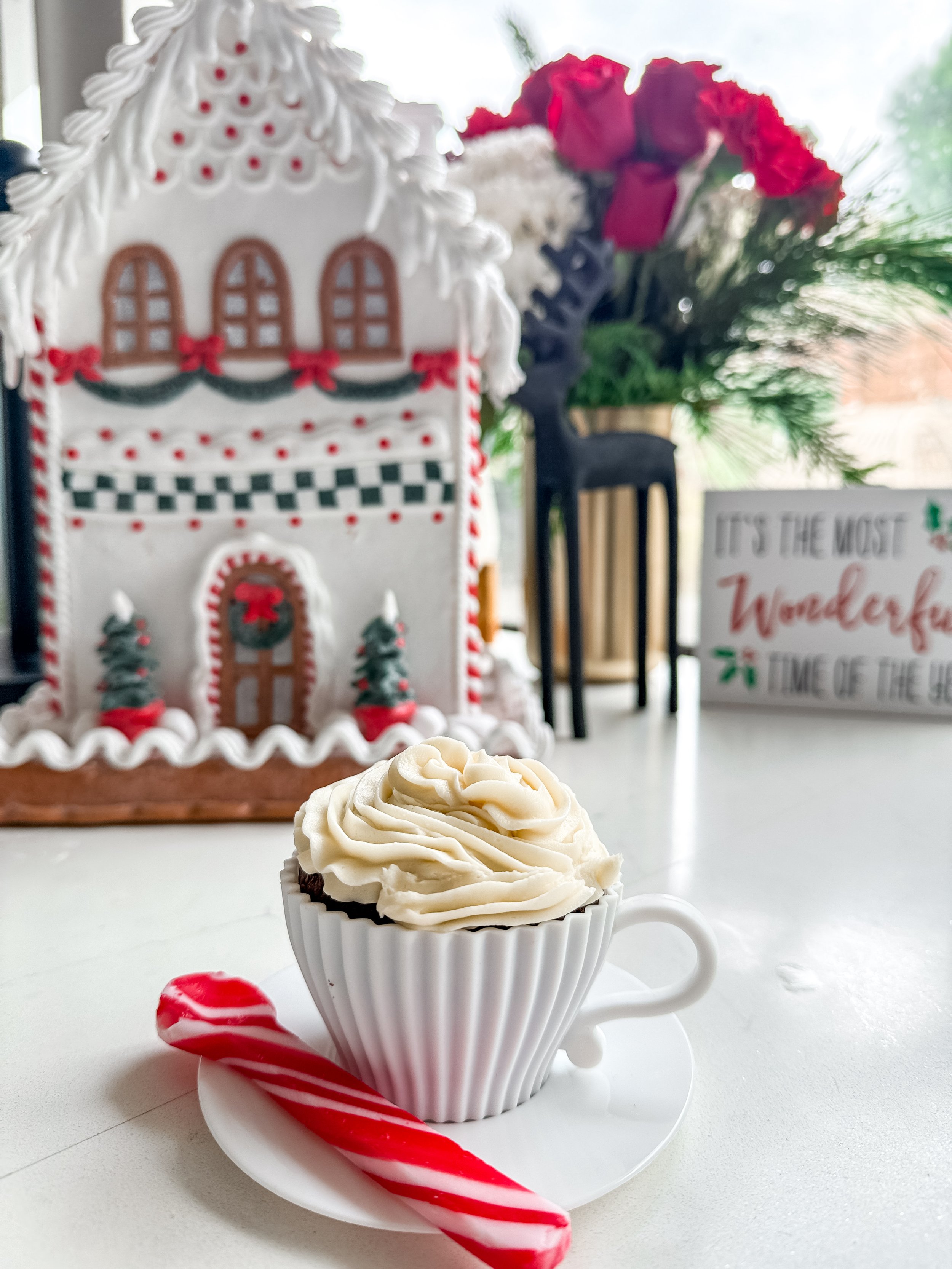 Cup of hot chocolate with whipped cream and a red and white candy cane in front, gingerbread house decoration, flowers, and holiday sign in the background.