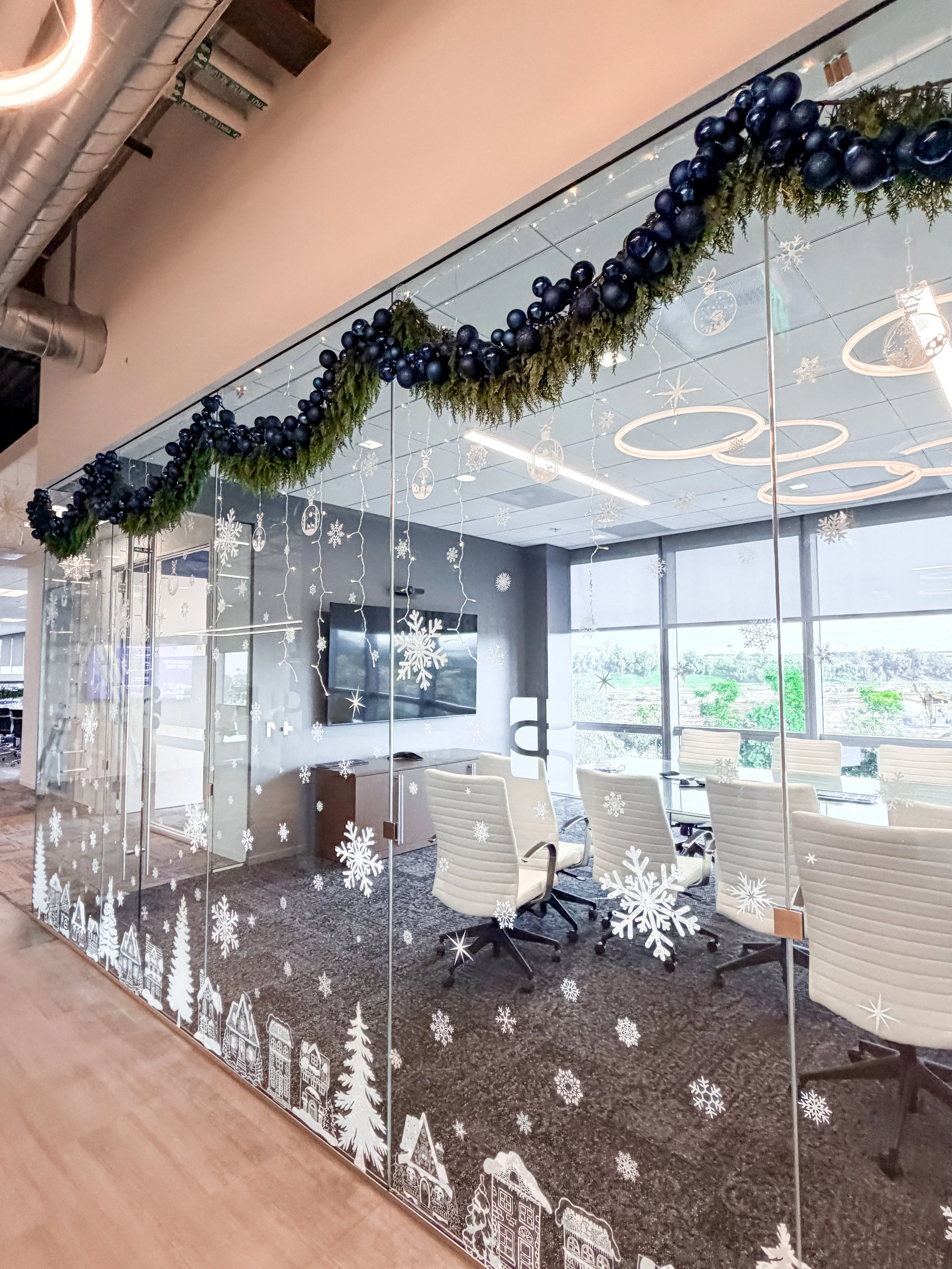Conference room with Christmas decorations, including a garland of pine and blue ornaments above the glass wall, and snowflake and winter village decals on the glass.