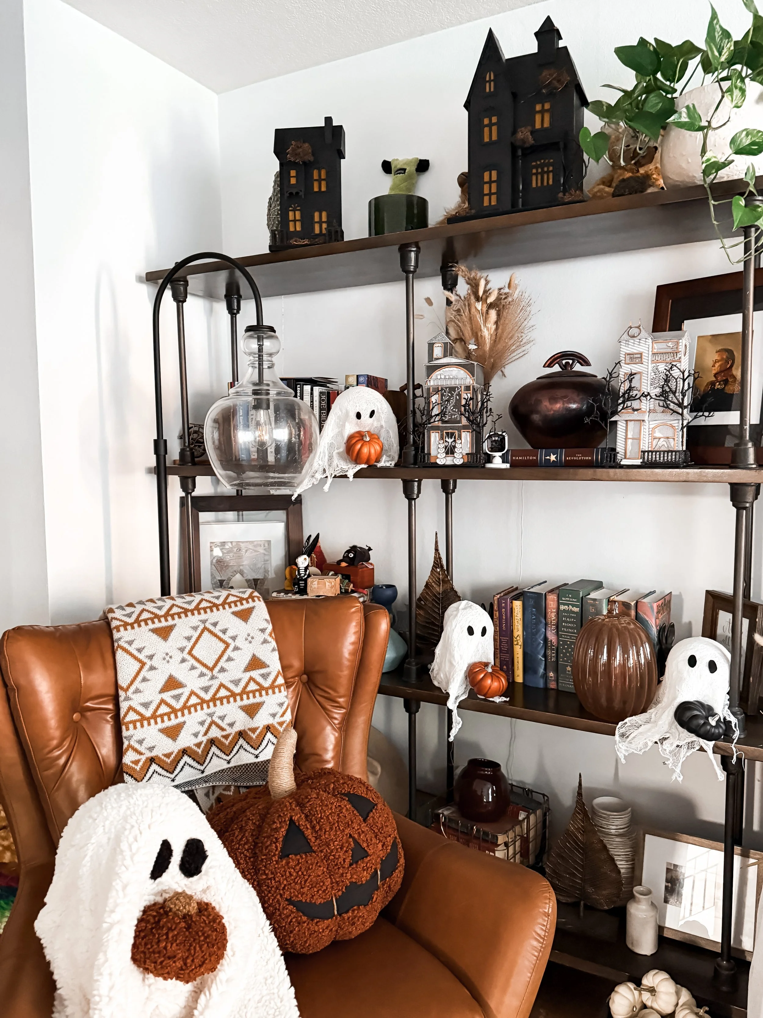 Halloween decorated living room shelf with ghost decorations, pumpkins, and spooky house models, and a leather chair with ghost and pumpkin plushies.