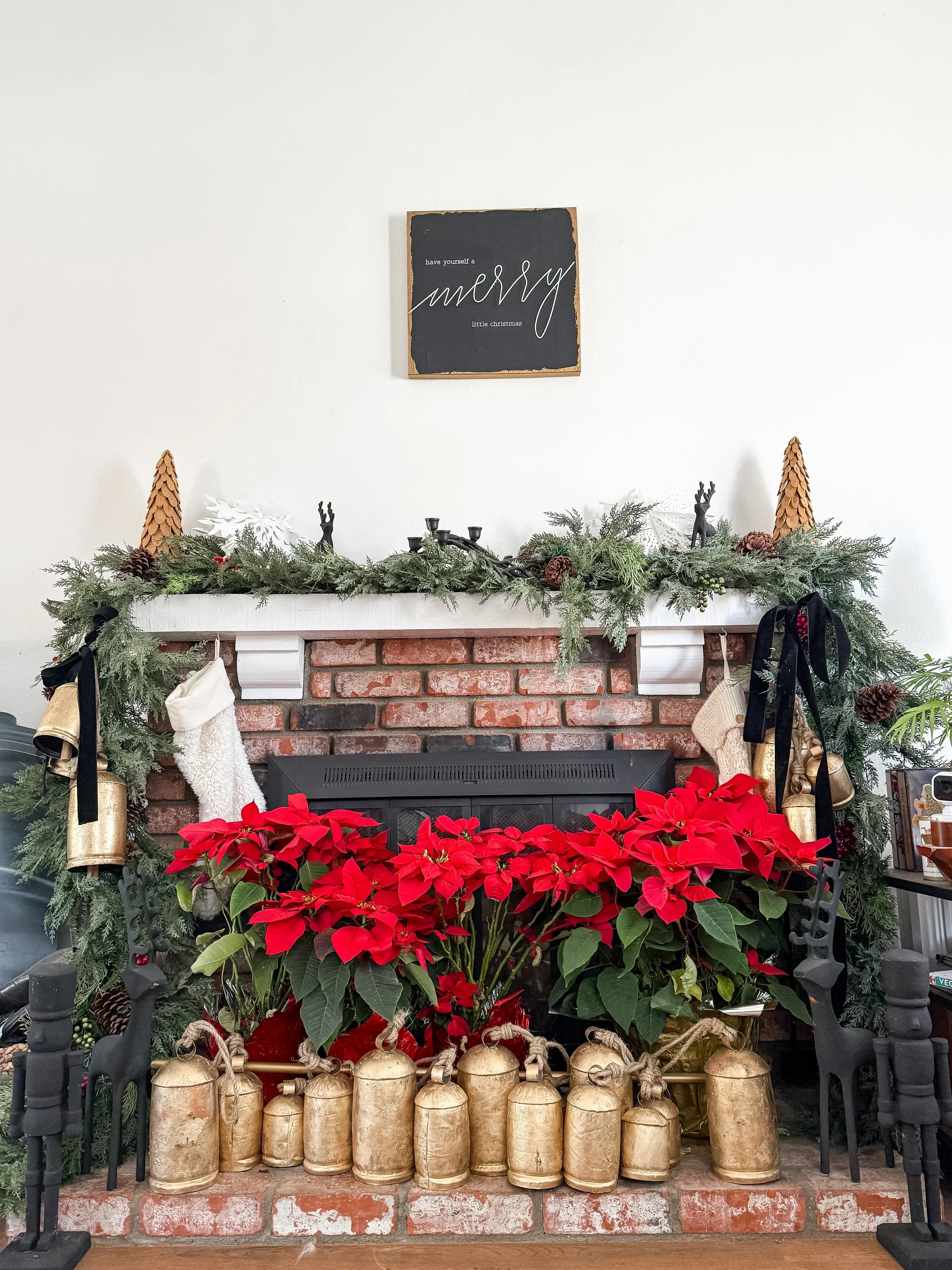 Decorated brick fireplace with greenery, gold bells, poinsettias, reindeer figures, and a blackboard sign on the wall that reads 'have yourself a merry little Christmas'.