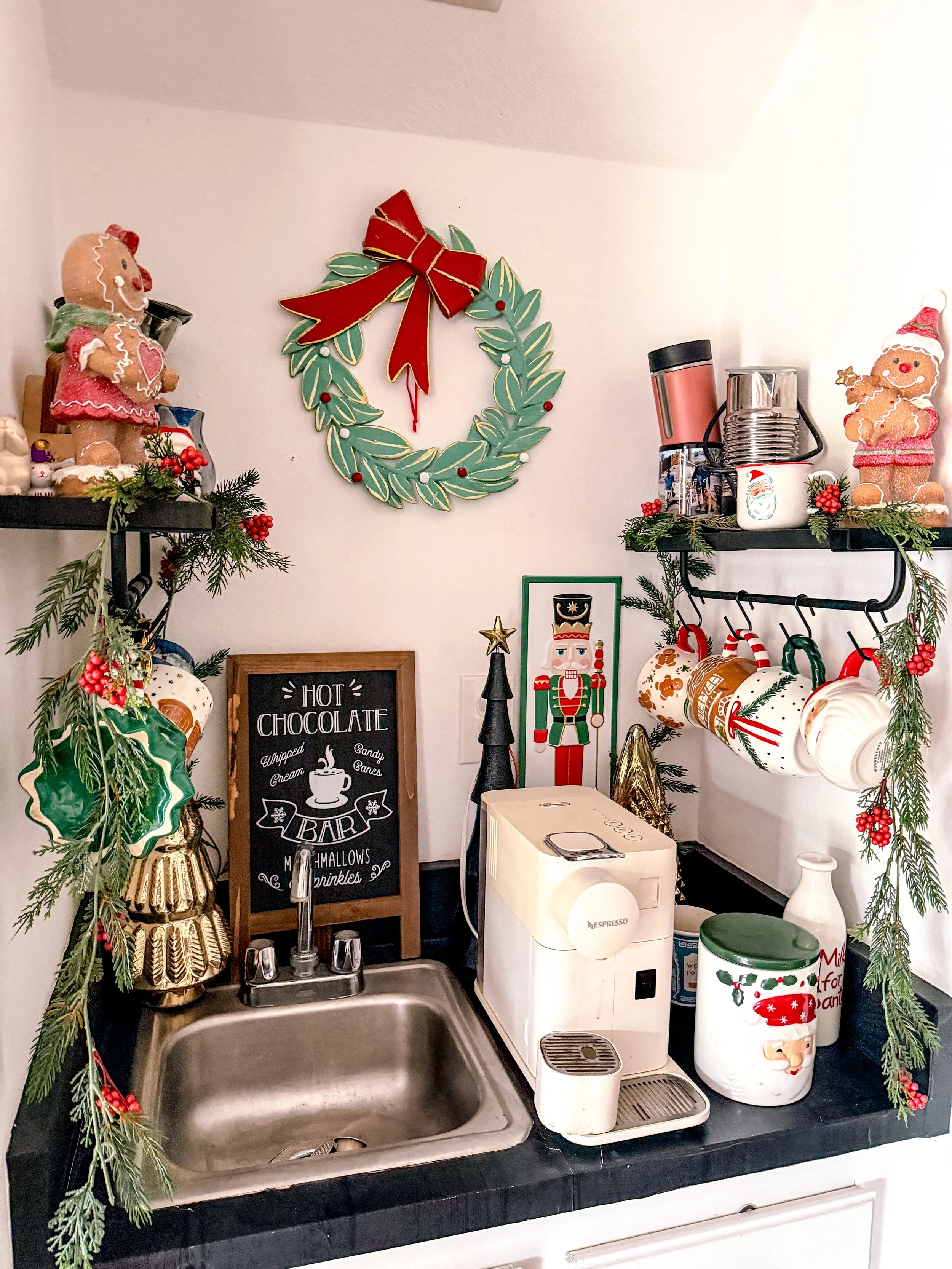 Christmas-themed kitchen corner with decorations, including a green wreath with a red bow on the wall, gingerbread cookies and holiday figurines on shelves, a coffee machine, a Santa-themed cookie jar, and festive garlands.