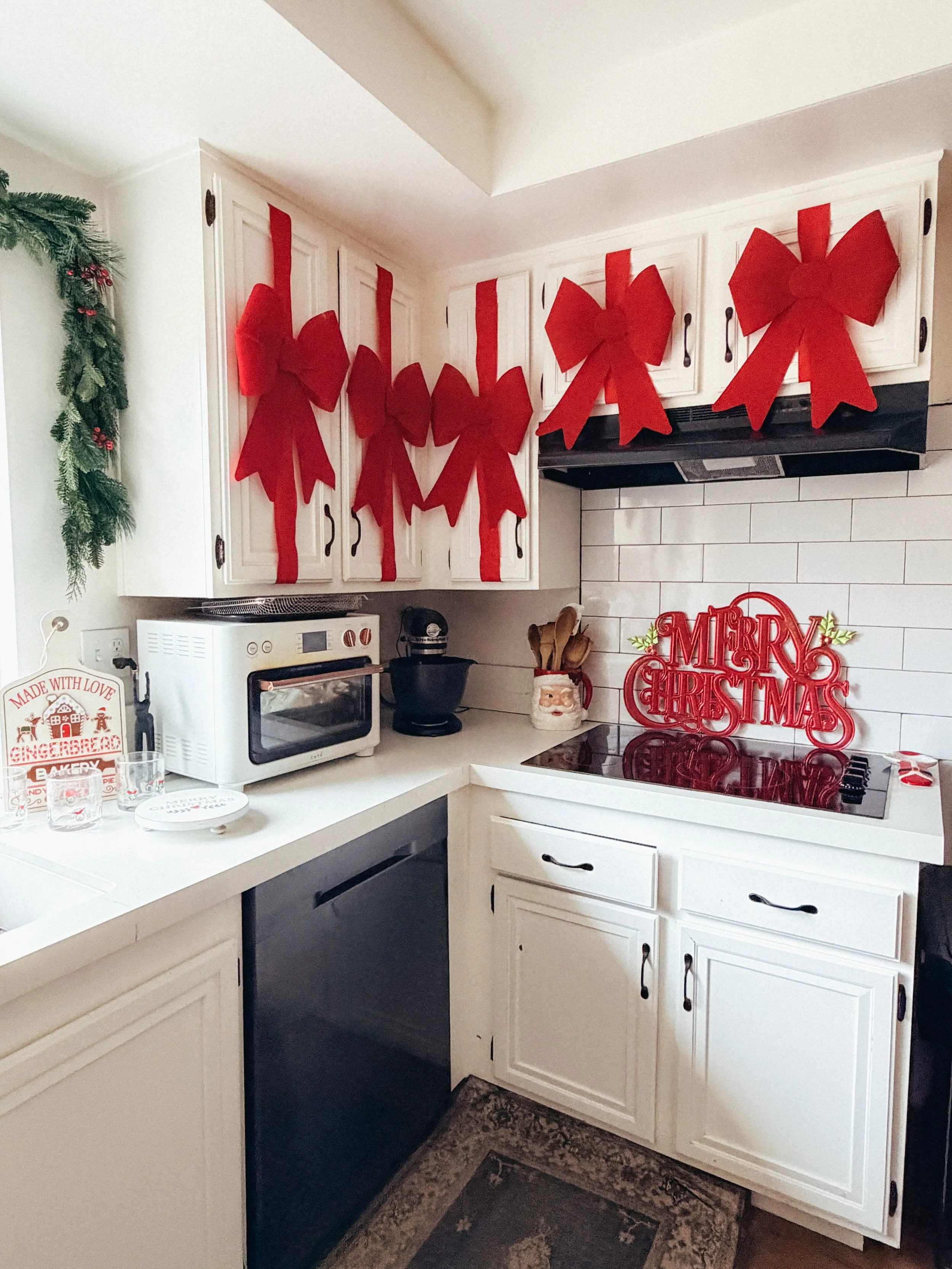 A Christmas decorated kitchen with white cabinets and a black stove. Red bows hang on the upper cabinets, and a red 'Merry Christmas' sign is on the stove. There are other Christmas decorations like a sign, glasses, and a wreath.