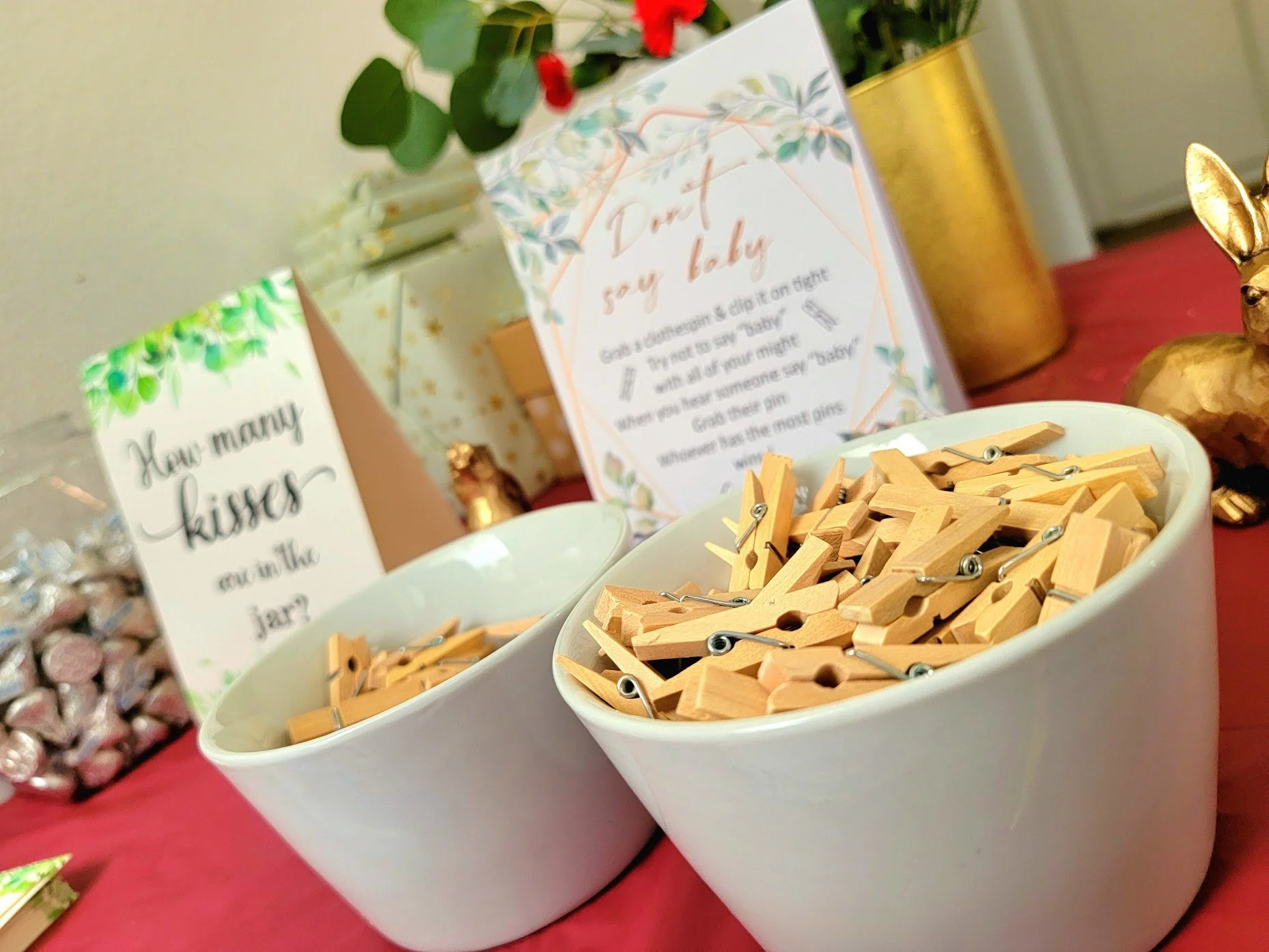 Two white bowls filled with small wooden clothespins on a red table, with colorful decorative signs and a plant in the background.