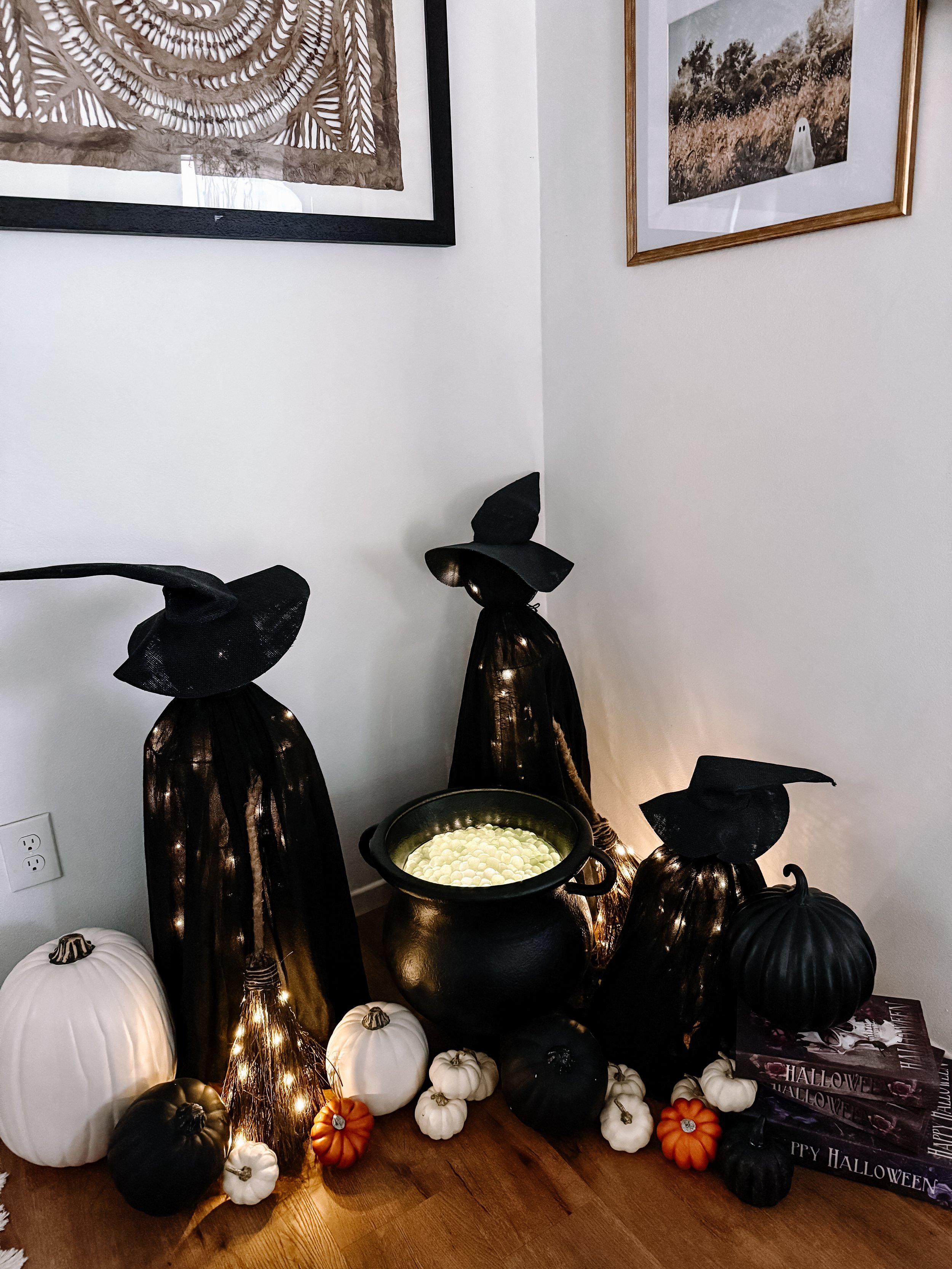 Decorative Halloween display with black witch hats on spooky black figures, white, black, and small orange pumpkins, a large black cauldron with a green bubbling liquid, string lights, and Halloween-themed books on a wooden surface at the corner of a
