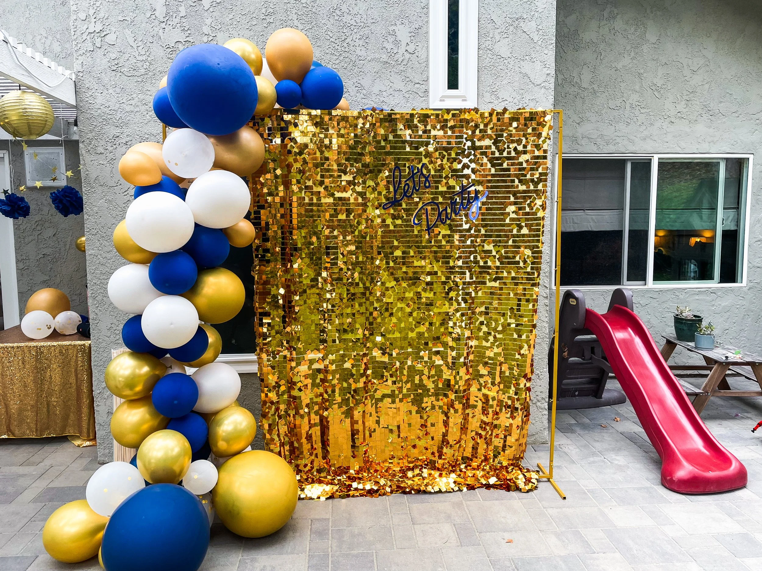 Decorative photo backdrop with gold, white, blue, and gold balloons arranged in an arch and on the ground. A shiny gold sequin backdrop with the words 'Let's Party' written on it in blue. A red slide and some potted plants on a picnic table to the ri