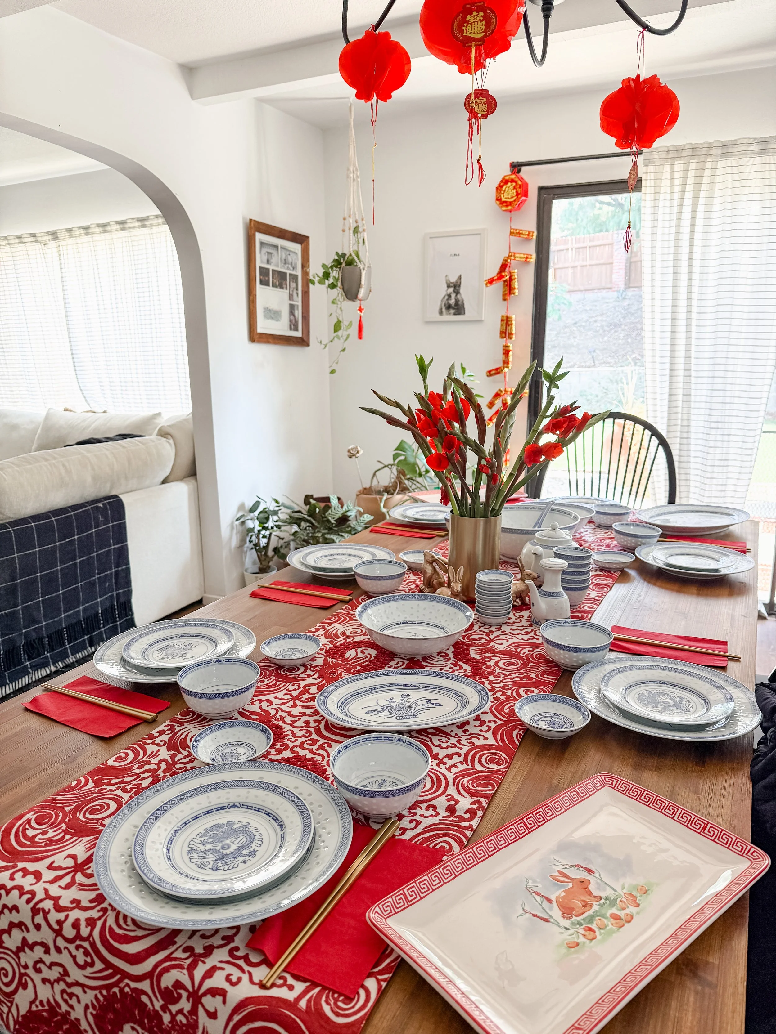 A dining table set with blue and white china and red napkins, decorated for a celebration with a red floral table runner and a vase of red flowers, in a bright room with hanging red lanterns and decorations.