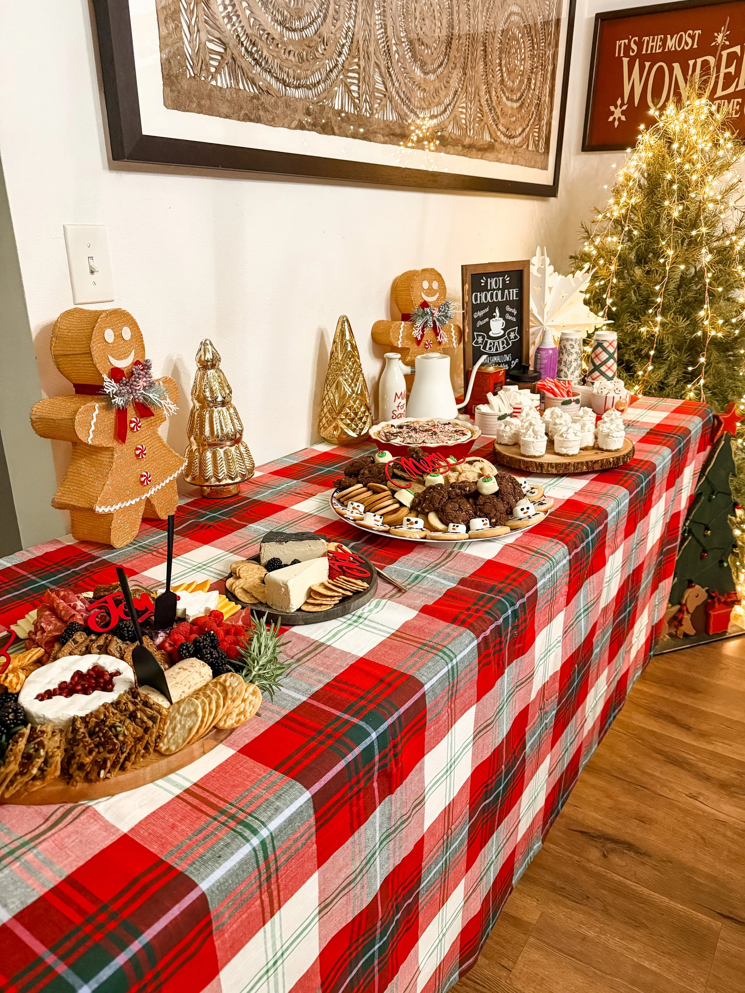 Christmas dessert table with gingerbread figures, cookies, chocolates, and cupcakes, decorated with holiday ornaments and a Christmas tree in the background.