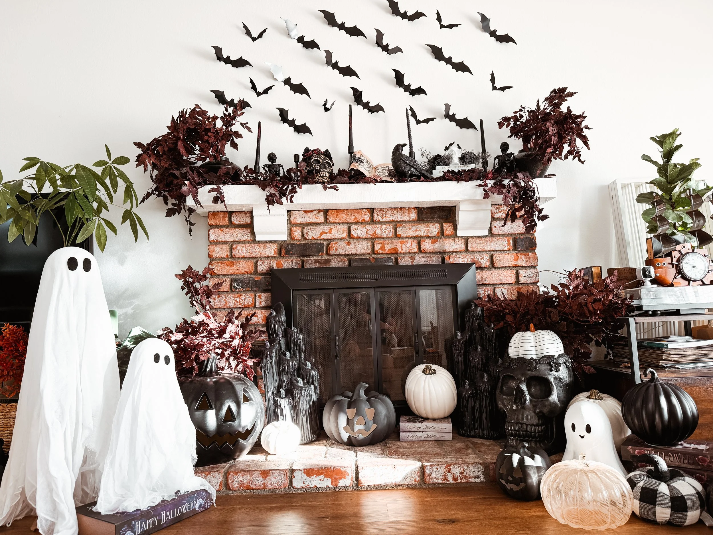 Decorated fireplace with Halloween-themed decorations including white ghost figures, black and white pumpkins, skulls, candles, bats, and dark foliage.