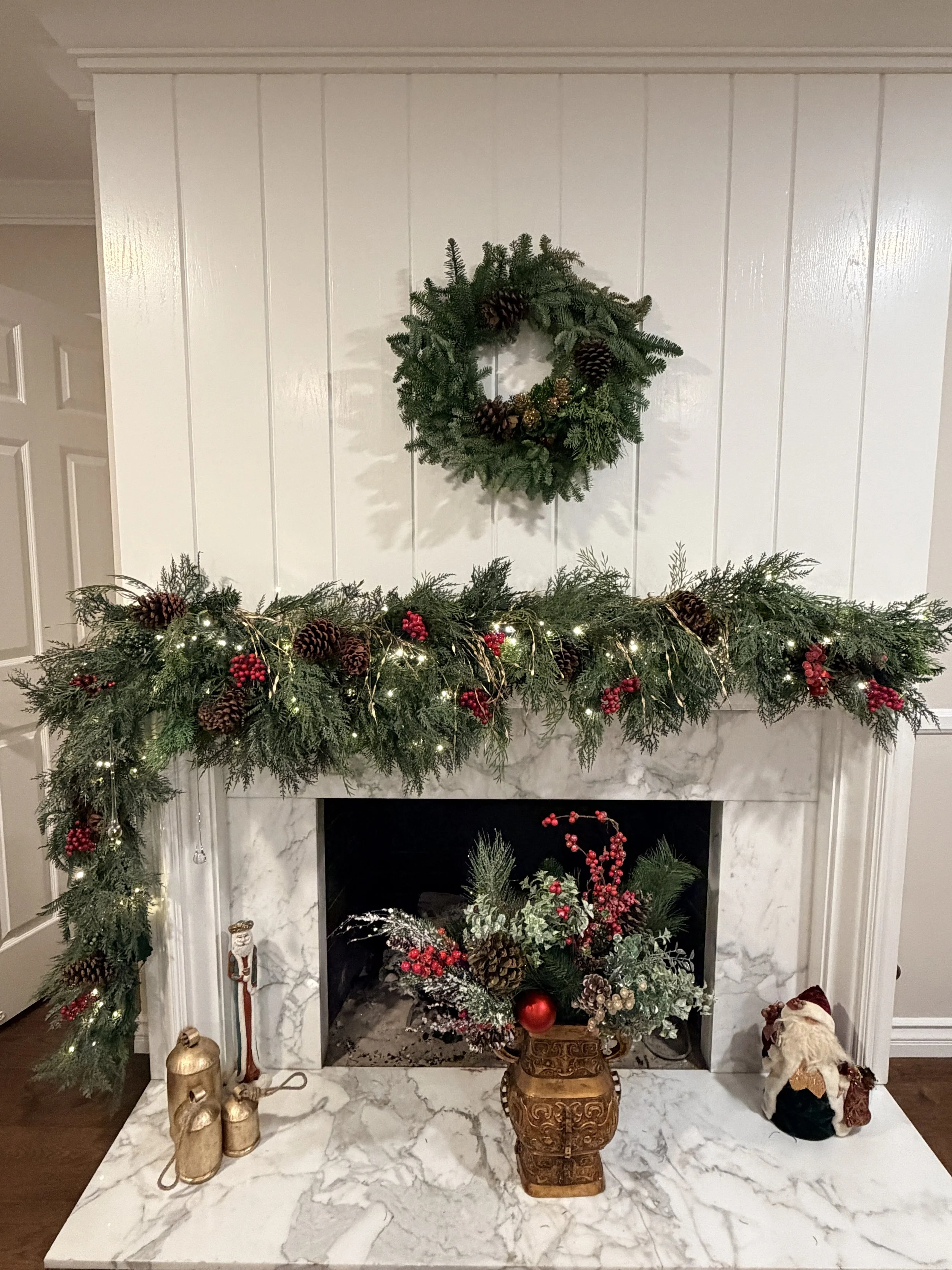 Decorated fireplace with Christmas greenery, pinecones, red berries, string lights, and holiday figurines.