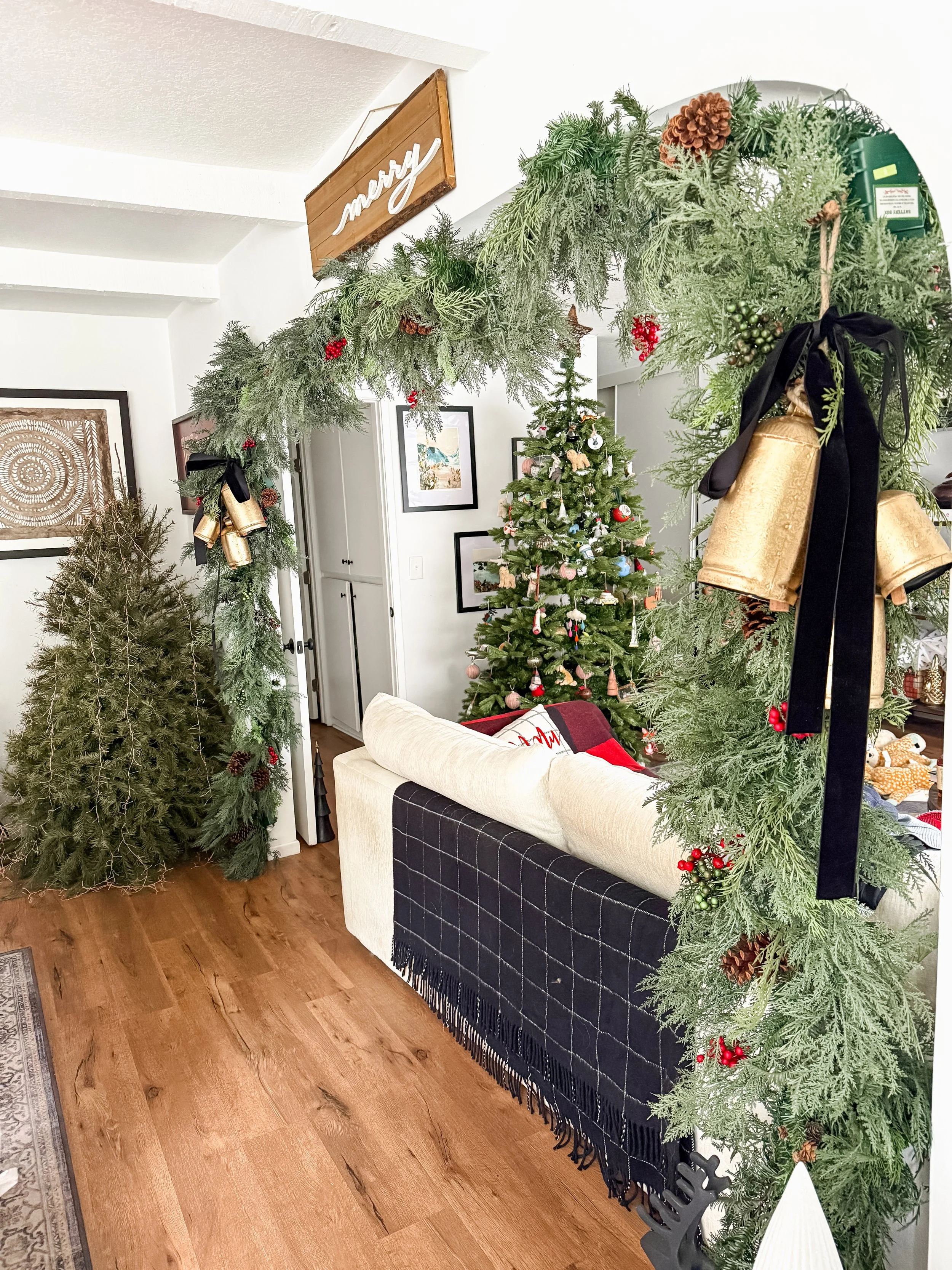 A decorated living room with a Christmas tree in the background, an archway decorated with greenery and holiday ornaments, including pine branches, red berries, and gold bells, with a wooden 'merry' sign hanging. There is a couch underneath the arch,