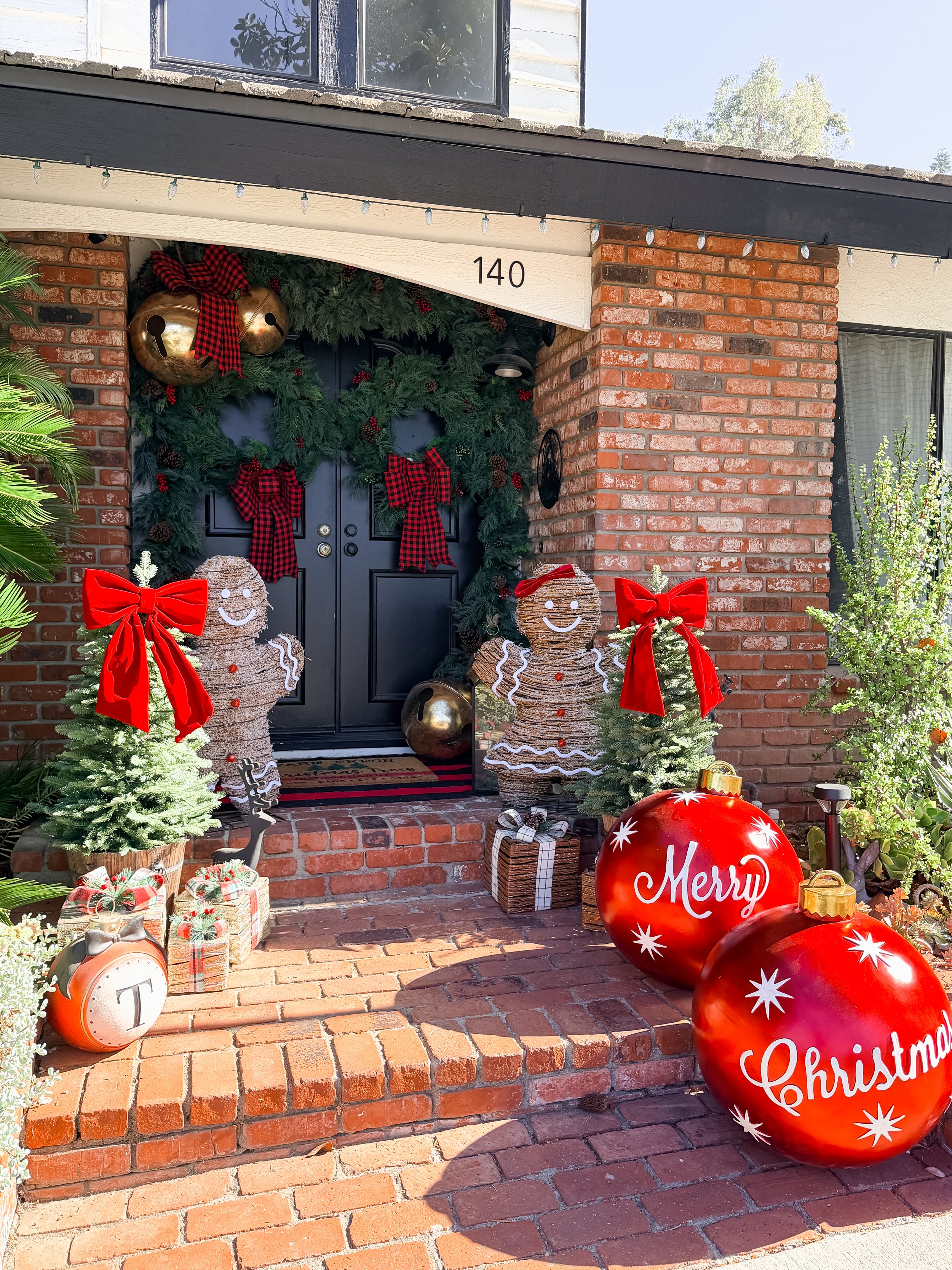 Festive Christmas porch with holiday decorations including red bows, gingerbread figures, large red ornaments with 'Merry Christmas' written, small potted Christmas trees, wrapped presents, and a large white bell with a red plaid ribbon above the doo