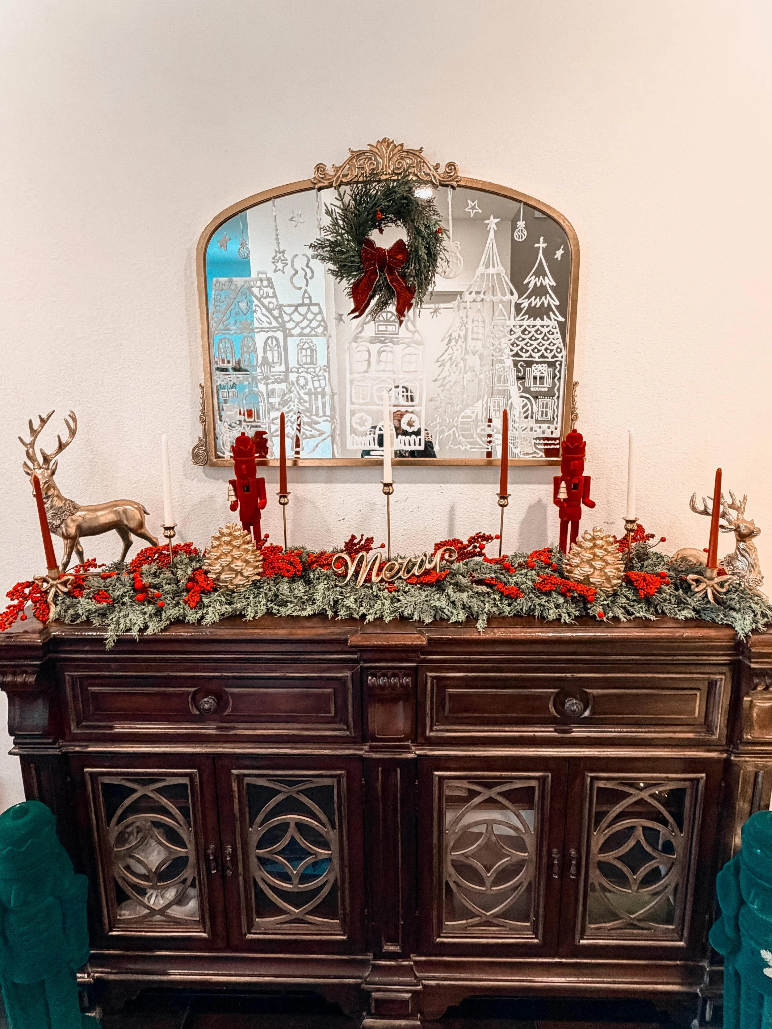Decorative brown wooden sideboard with Christmas and winter holiday decor, including red and gold reindeer figurines, pinecones, candles, red berries, and a 'New Year' sign, with a wall mirror above reflecting a snowy winter scene drawing.