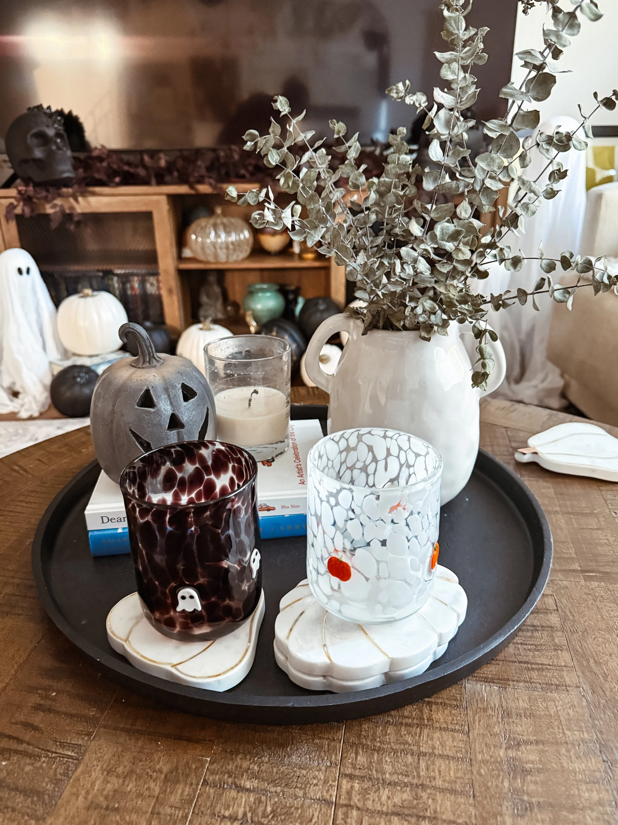 Halloween-themed table centerpiece with a ceramic ghost, a black pumpkin lantern, a glass candle holder, and decorated drinking glasses on marble coasters, all on a black tray, with a vase of dried eucalyptus on a wooden table.