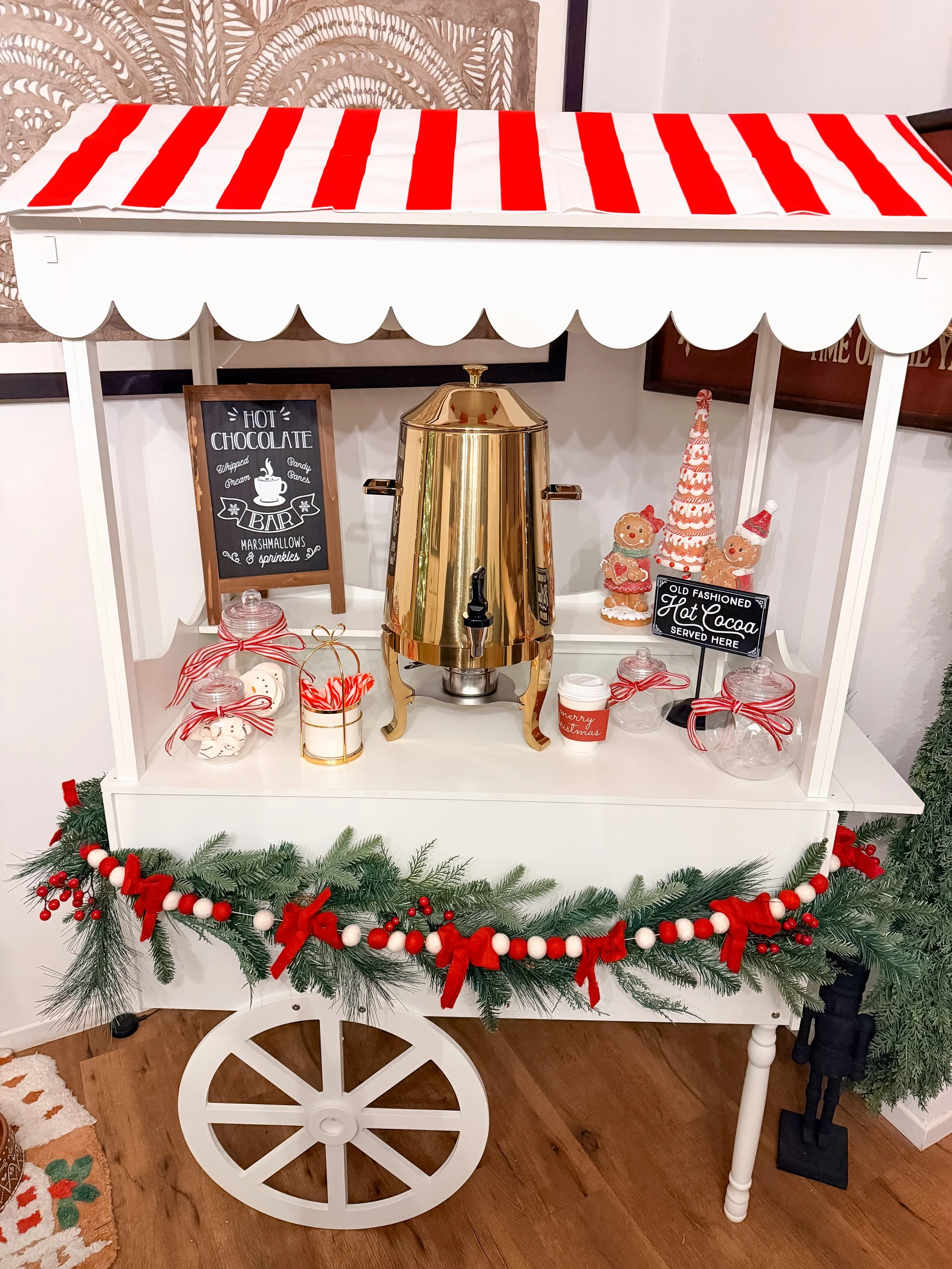 Christmas hot chocolate stand decorated with red and white bows, pine garland, and holiday-themed figurines, with a gold hot chocolate dispenser and jars of marshmallows.