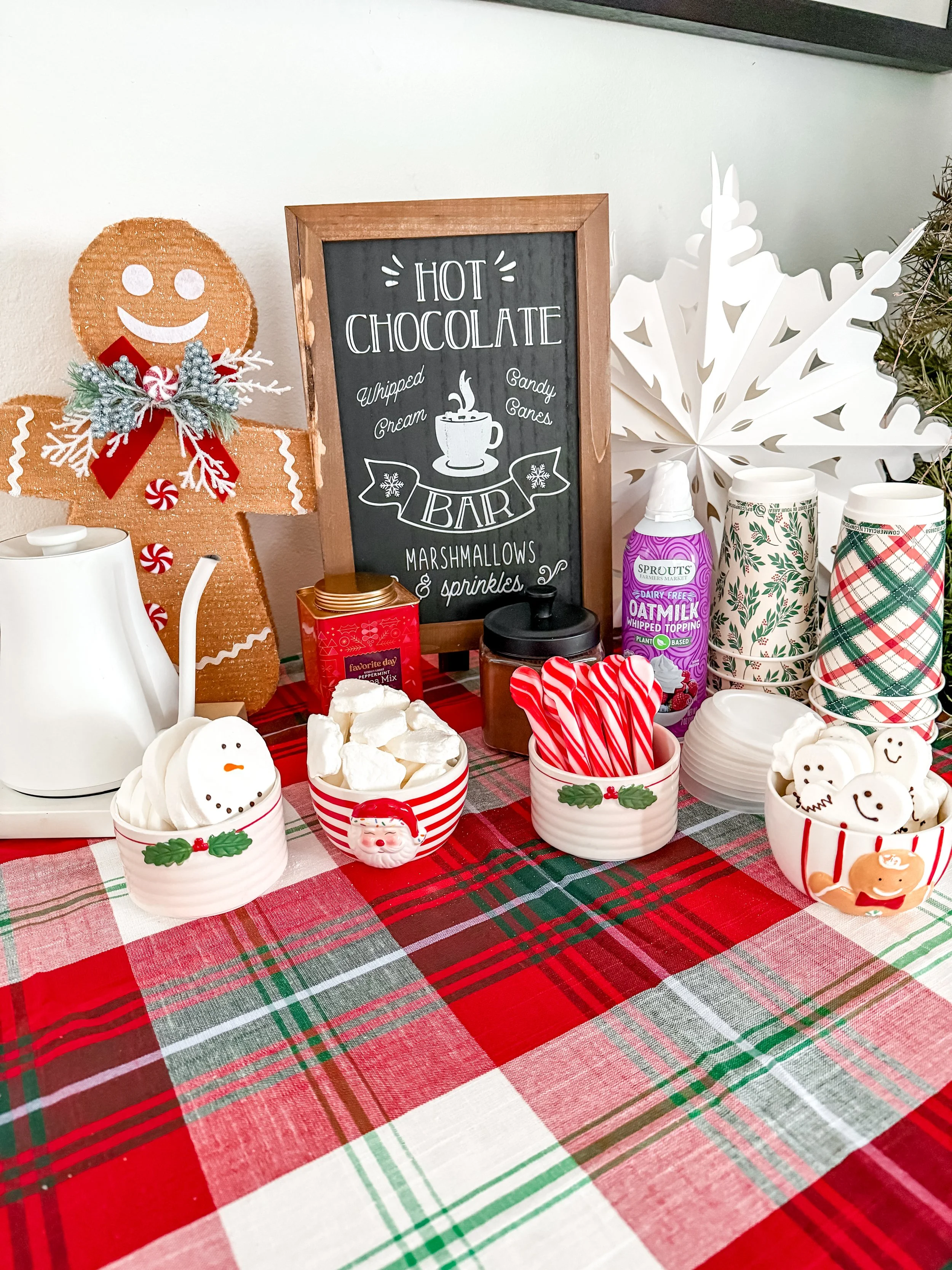 Festive holiday table with marshmallow treats, candy canes, and seasonal decorations including a gingerbread figure, a snowman, and a Santa, with a chalkboard sign for a hot chocolate bar.