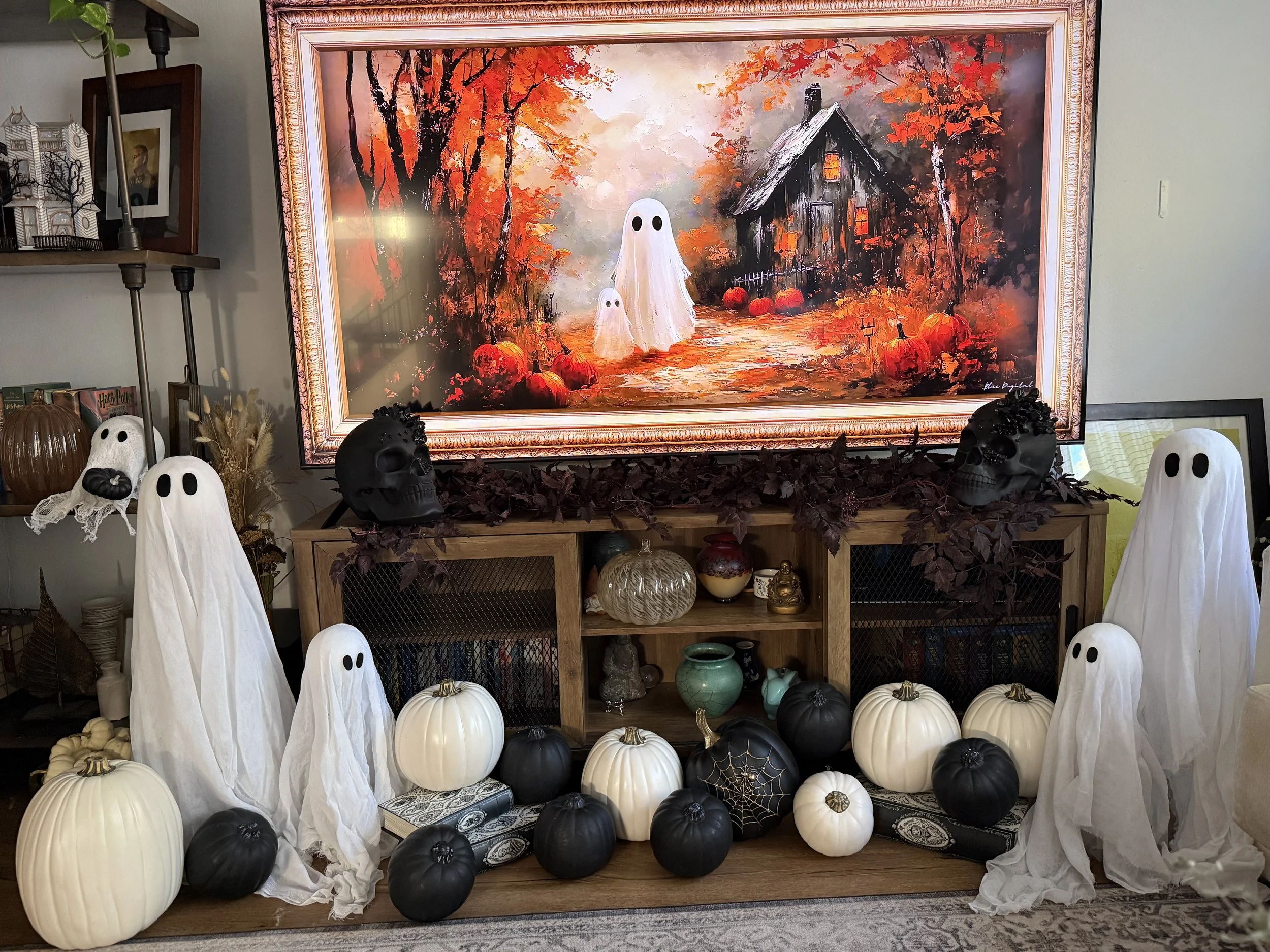 Halloween and autumn decorations in front of a painting of a spooky ghostly scene with pumpkins, ghosts, and a haunted house.