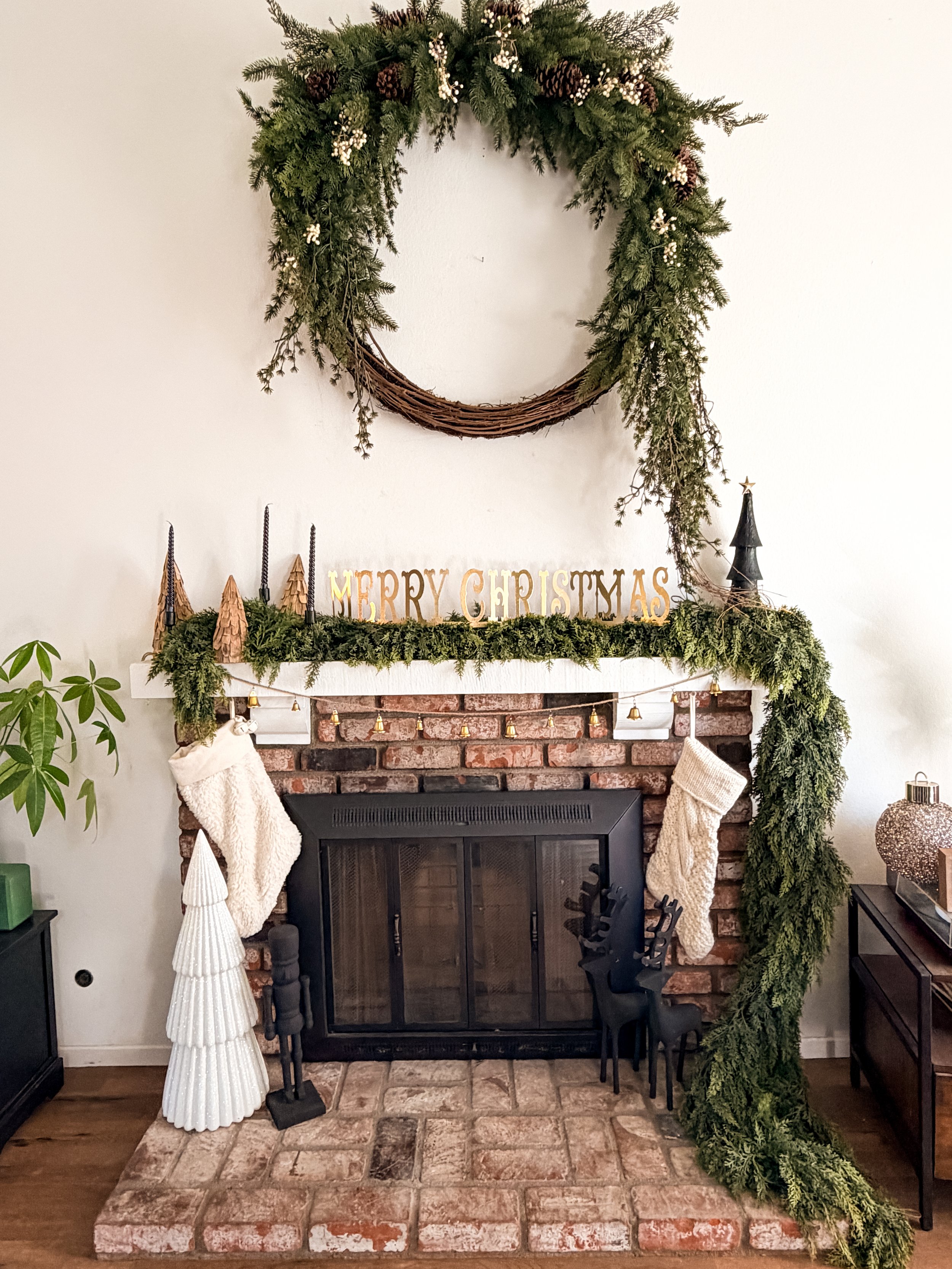 Decorated brick fireplace with Christmas stockings, garland, and a "Merry Christmas" sign on the mantle. A large green wreath hangs above, and there are Christmas figurines including reindeer and tree-shaped decorations.
