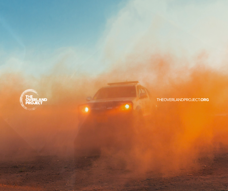 A vehicle driving through a cloud of orange dust in an outdoor desert landscape with a blue sky