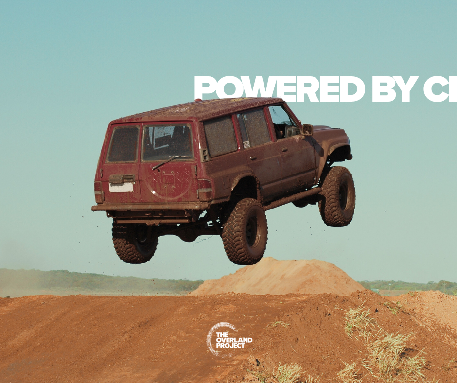 A muddy, red, four-wheel-drive vehicle jumps over a dirt mound in an off-road setting with a pale blue sky in the background. Text overlay reading 'POWERED BY CH' and a logo that says 'THE OVERLAND PROJECT'.