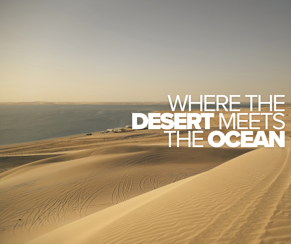 Desert dunes meeting the ocean with a city and boats in the background