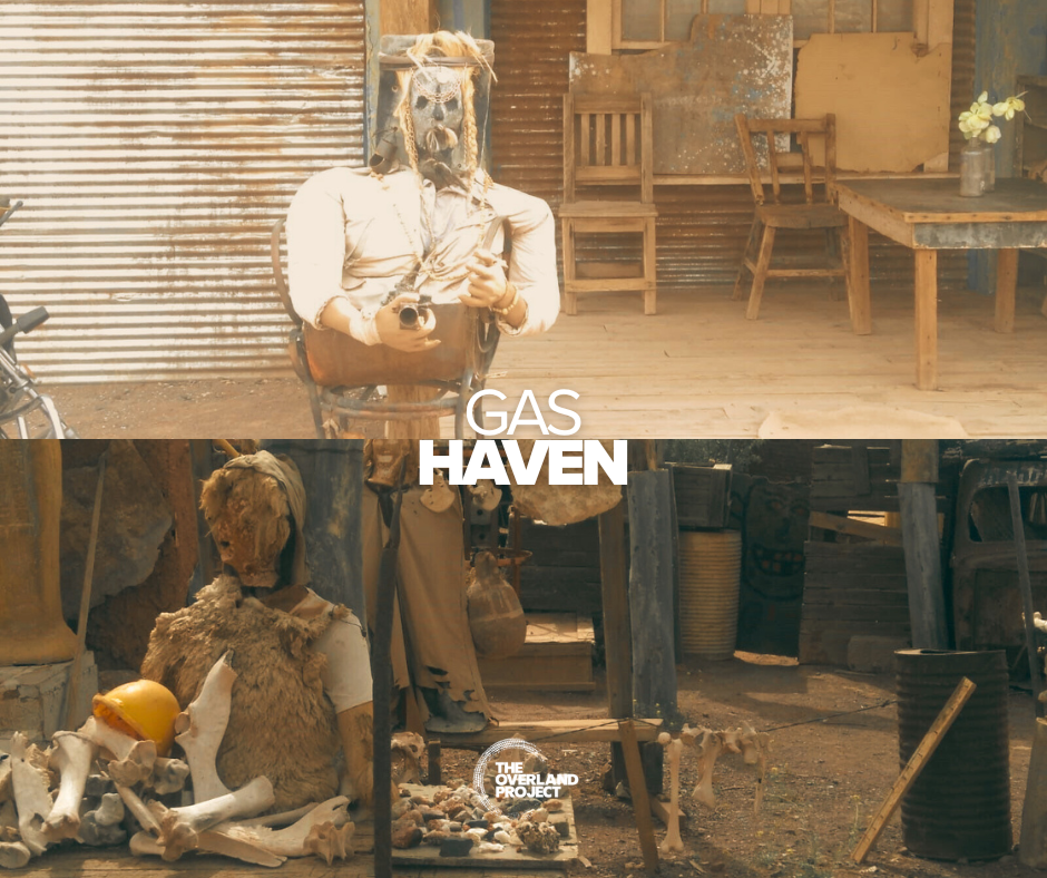 A person in a mask and costume taking a selfie with a camera inside a rustic setting with chairs and tables, and a displayed skeleton with a yellow helmet and animal fur. Text overlays say 'GAS HAVEN' and 'THE OVERLAND PROJECT.'