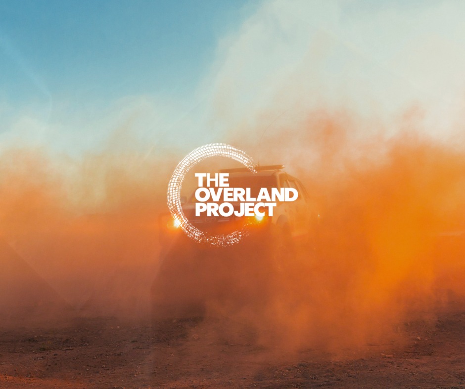 A vehicle emitting orange smoke or dust, with the logo and text "The Overland Project" overlayed on the image.