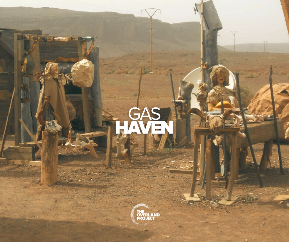 Desolate desert landscape with makeshift wooden structures, animal skulls, and skeletal remains arranged as decor or sculptures, under power lines. Text overlay reads 'Gas Haven' with 'The Overland Project' logo at the bottom.