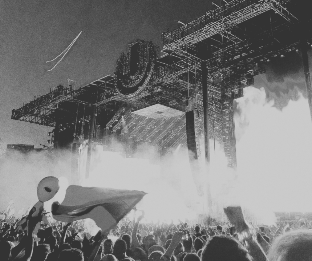 A large outdoor concert stage with bright lights, fog, and a crowd of people holding up flags and signs.