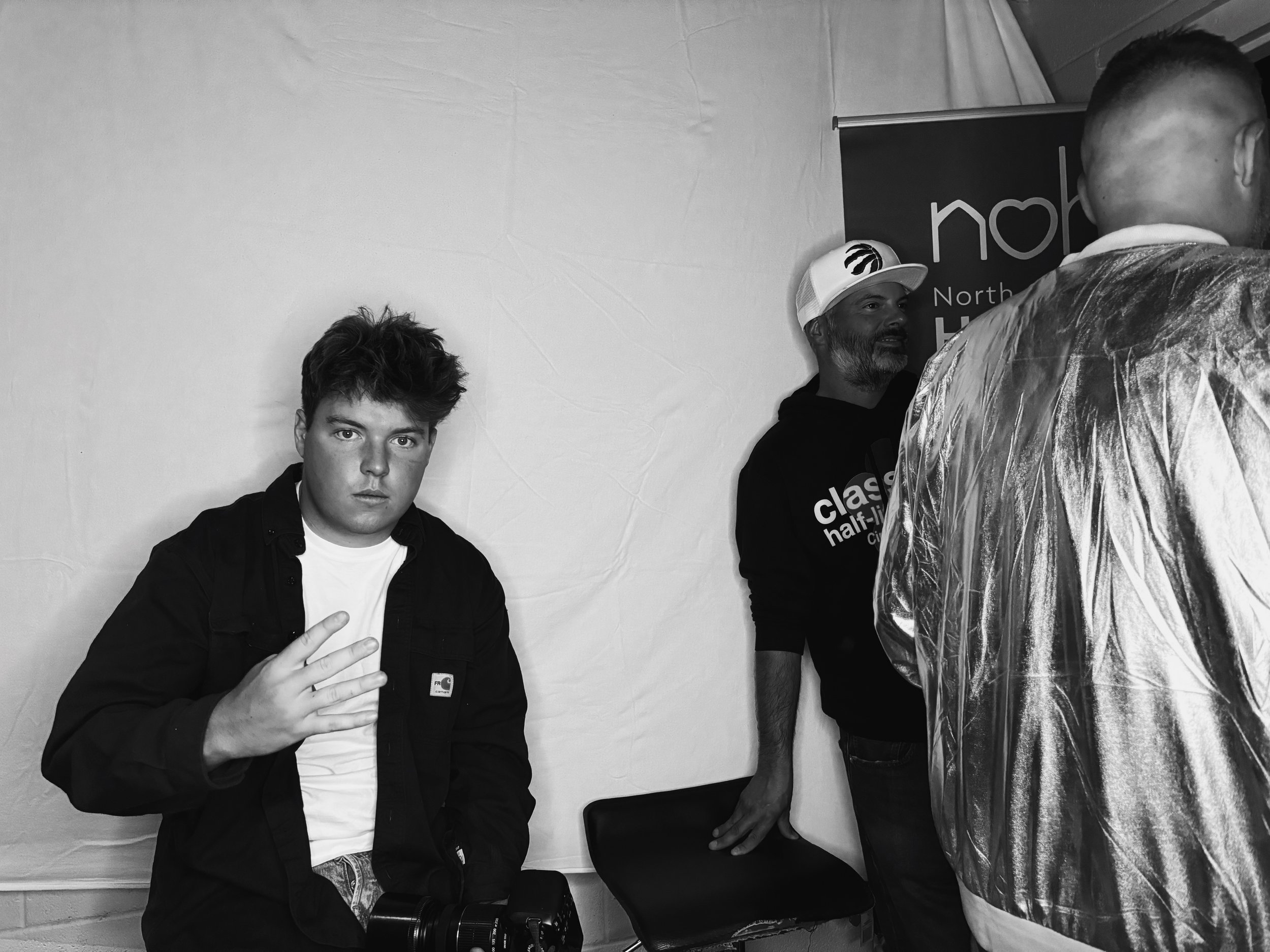 A black and white photo of three men standing indoors against a plain wall. The man on the left has dark hair and is making a peace sign. The man in the middle has a beard, is wearing a cap, and is smiling slightly. The third person on the right has short hair and is wearing a plastic poncho, facing away from the camera. Part of a banner with the text 'north' is visible behind them.