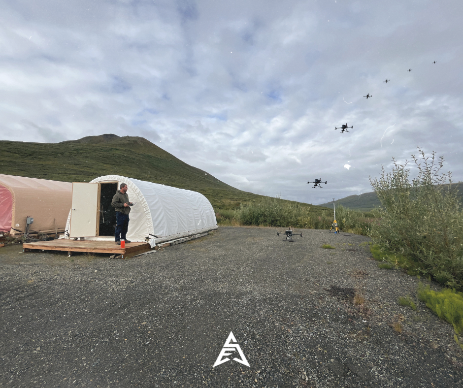 A person standing outside a white tent on a gravel surface, operating a remote control. Multiple drones are flying in the sky against a backdrop of mountains and cloudy weather.