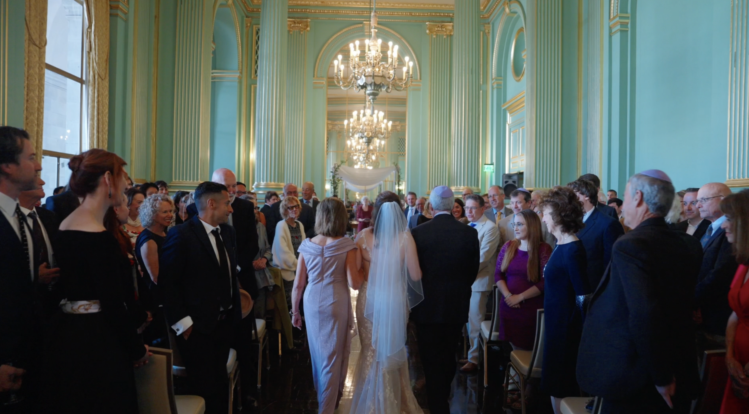 A san francisco wedding ceremony with a bride and groom standing in front of a large gathering of guests in a decorated hall with tall green walls and chandeliers.