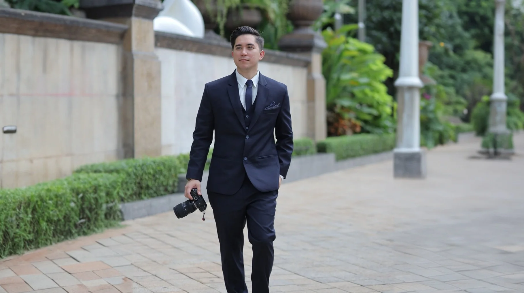 David (davideographer) in a dark suit and tie walking outdoors on a paved pathway, holding a camera in his right hand, with greenery and lamp posts in the background.