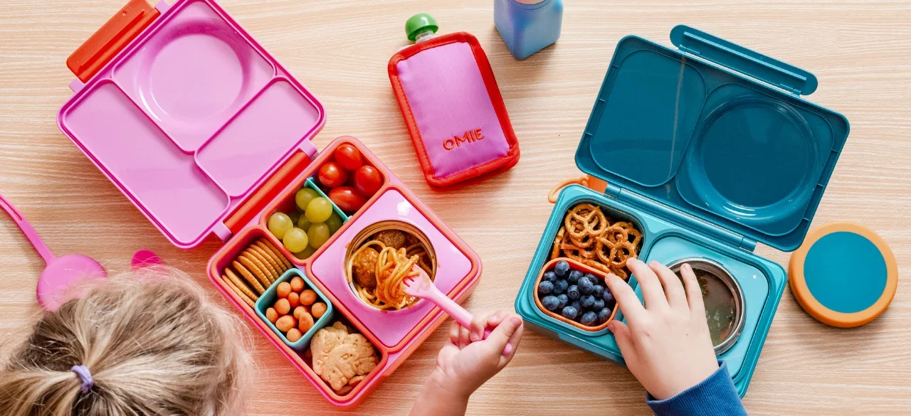 Children's lunchboxes on a table with yogurt, blueberries, pretzels, crackers, grapes, and other snacks, along with a pink spoon, a small pink container, and a blue water bottle.