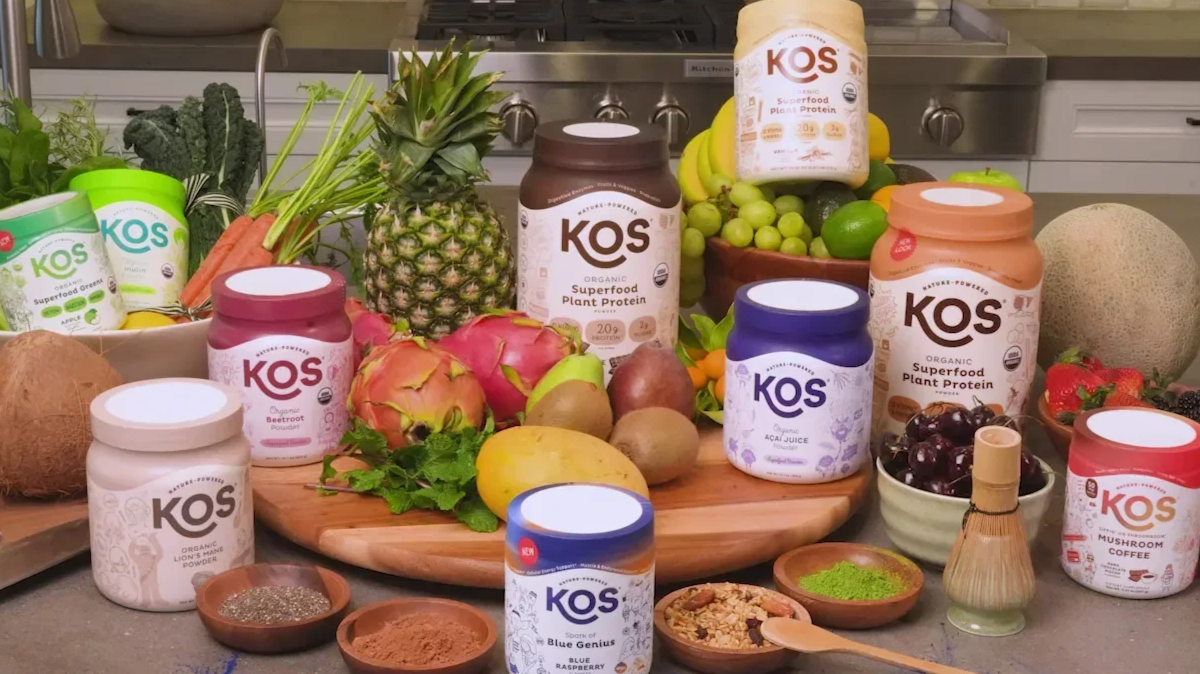 A variety of KOS organic superfood supplements and fresh fruits and vegetables displayed on a kitchen counter, including pineapple, dragon fruit, grapes, cucumbers, carrots, a cantaloupe, strawberries, blackberries, and bowls of powders and seeds.