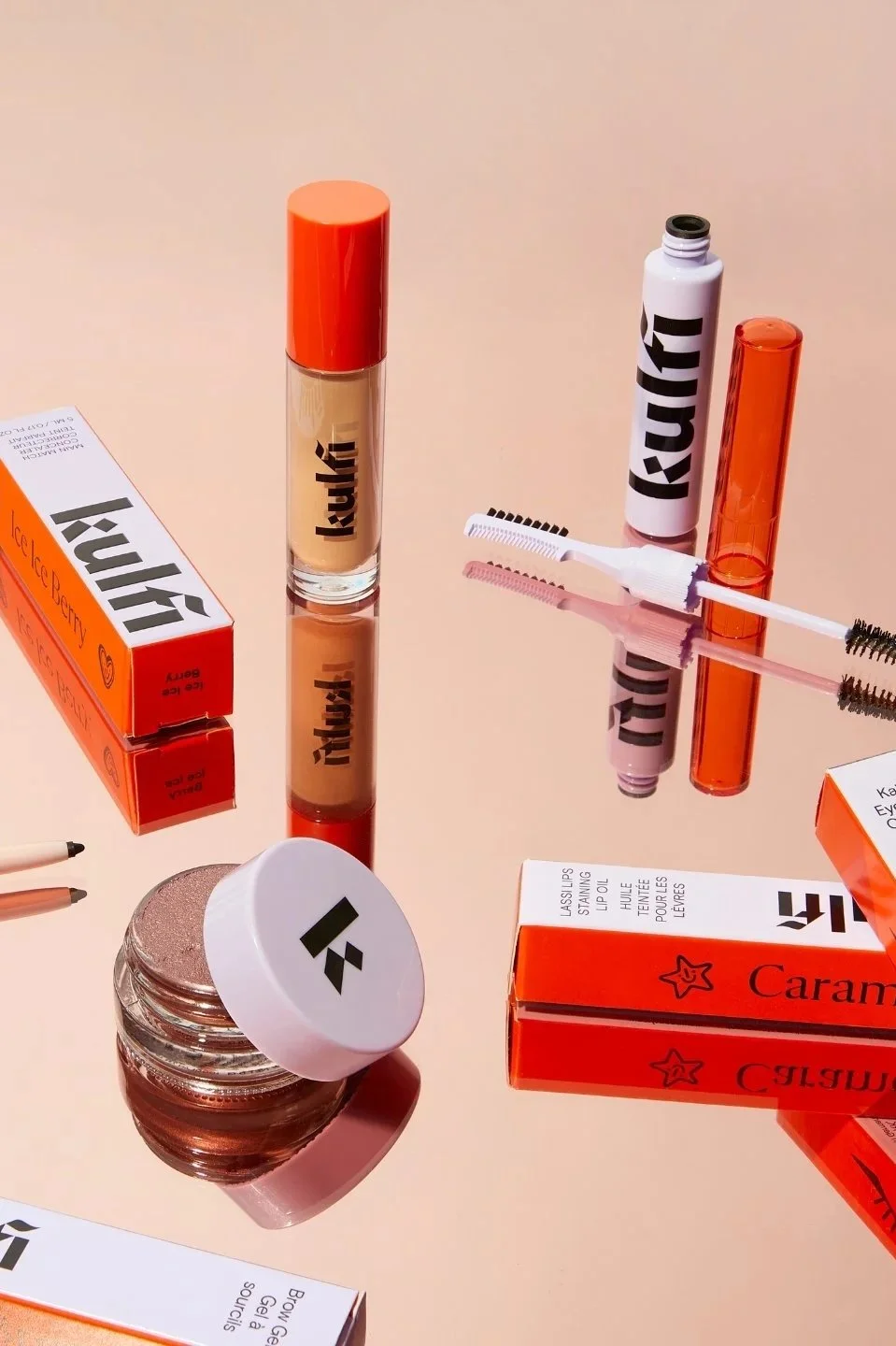 Assorted Kaja beauty products including lip gloss, mascara, brow gel, and boxed eye makeup on a reflective surface.