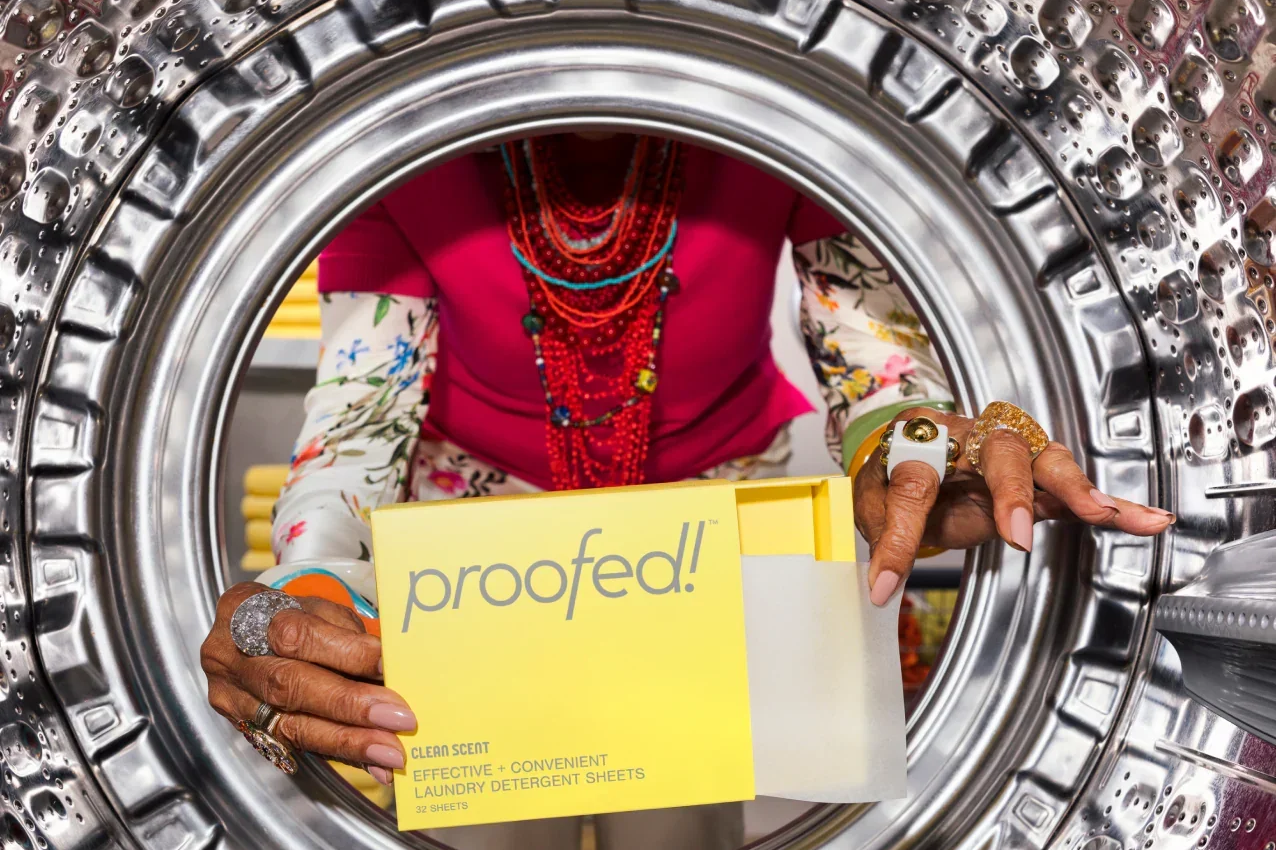 Person loading a washing machine, seen through the door, holding a yellow box of 'proofed!' laundry detergent sheets.