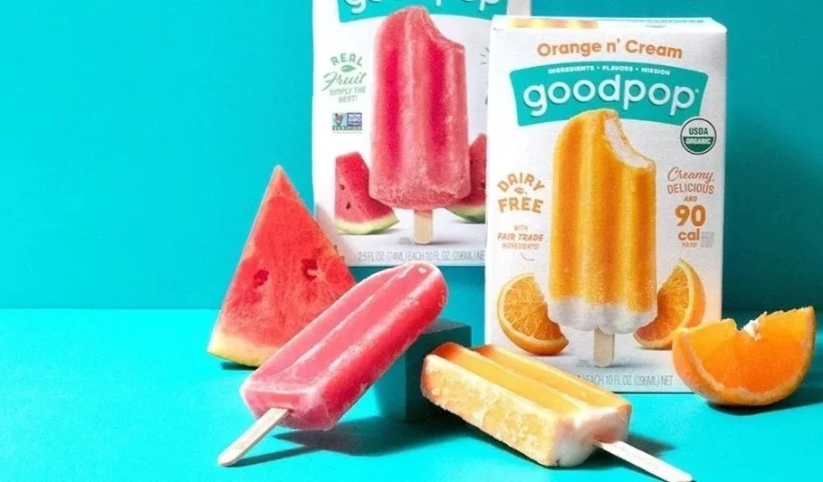 Assorted fruit popsicles and juice boxes on a blue surface with a turquoise background. There are watermelon, orange, and lemon flavored popsicles, along with slices of watermelon, orange, and lemon beside the popsicles.