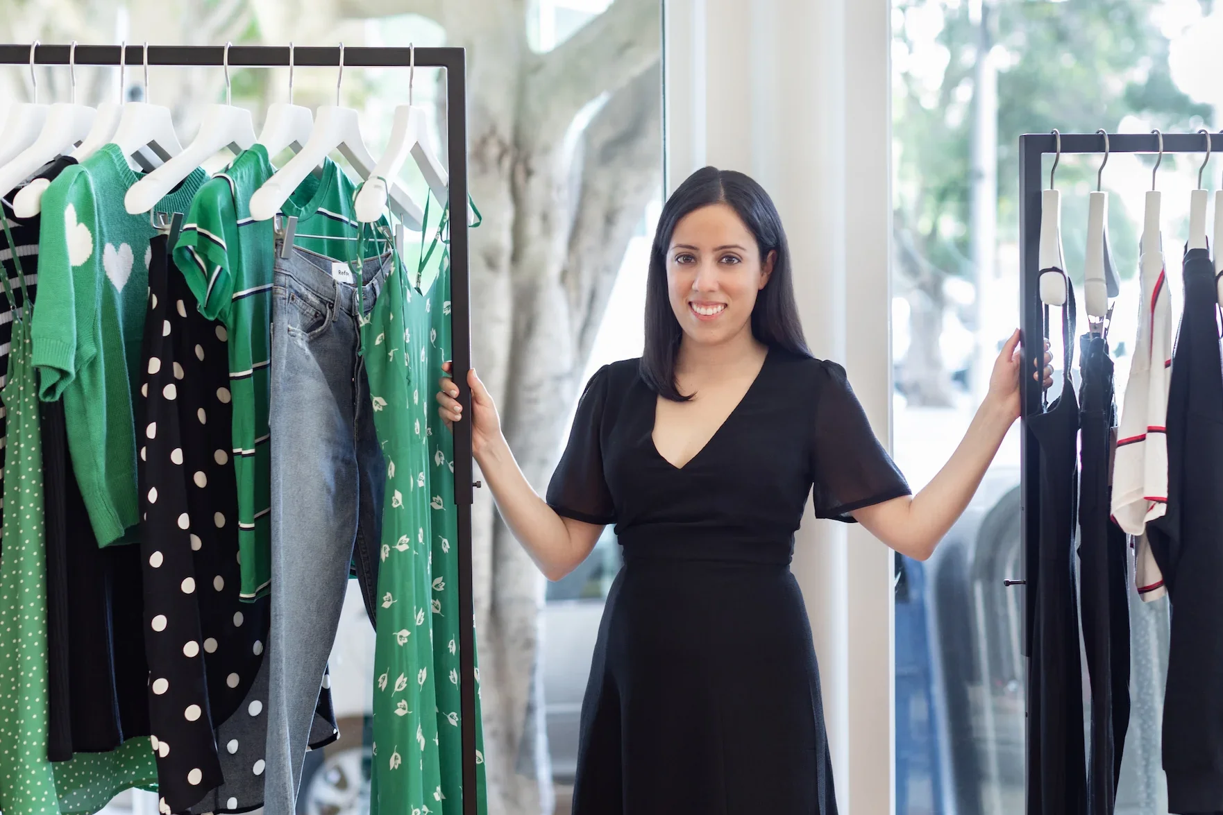 A woman smiling and shopping for clothes in a store, holding clothes racks with green, black, and white patterned dresses and a pair of jeans.
