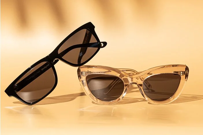 Two pairs of sunglasses, one with black frames and dark lenses, the other with clear frames and dark lenses, on a beige surface.