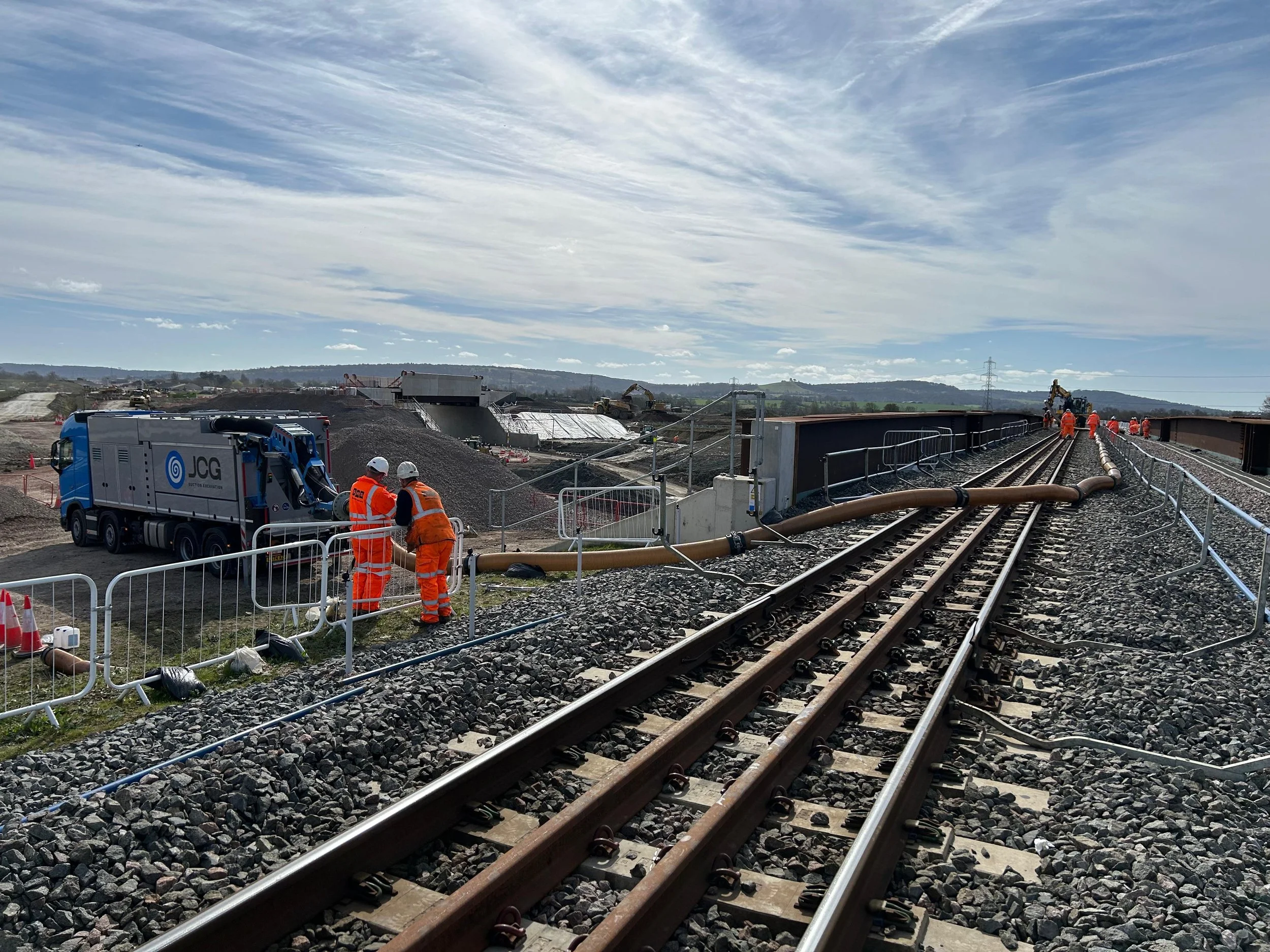 Construction workers in orange safety gear working on railway tracks on a bridge during daytime.
