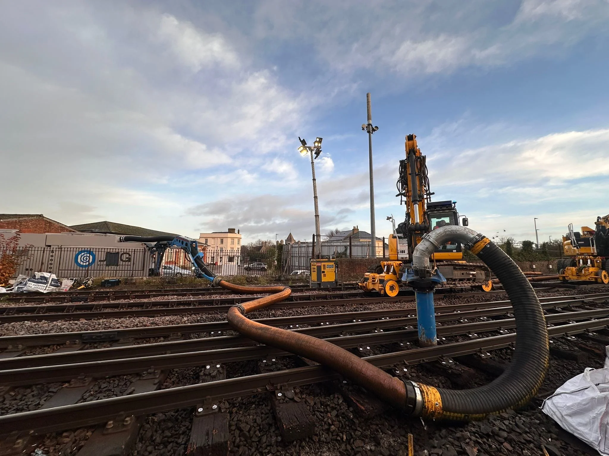 Construction equipment on railway tracks, including large hoses and machinery, with a fence and buildings in the background on a cloudy day.