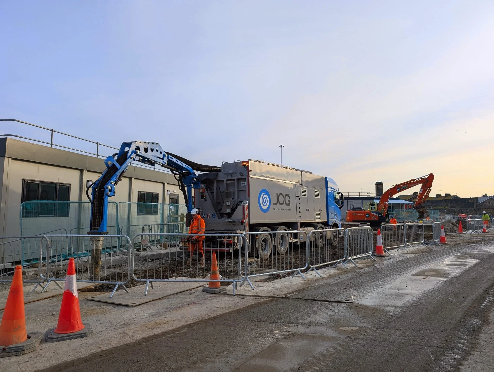 Construction site with heavy machinery and workers in safety gear, orange cones, and metal barriers, with a cloudy sky in the background.