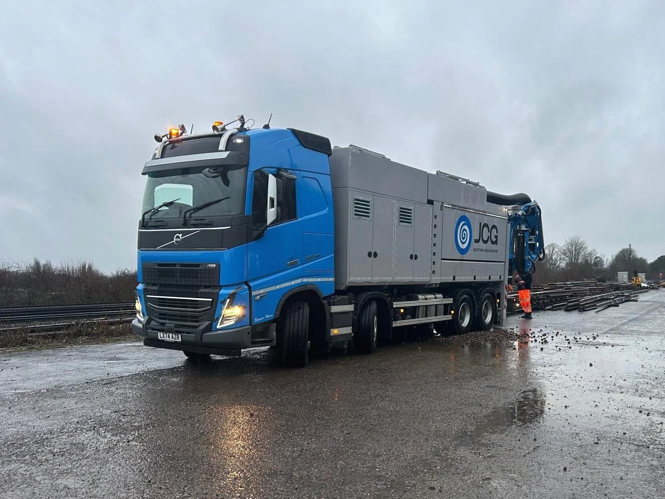 A large blue and gray JCG section excavator truck parked on a wet road near railway tracks, with a worker standing nearby.