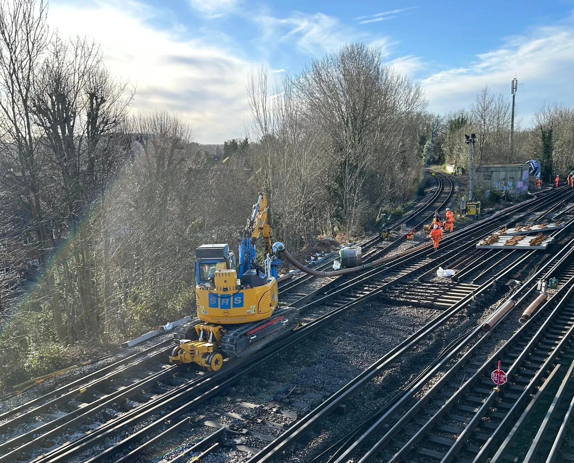 Construction workers in orange safety gear repairing train tracks on a railway. A small excavator is using a hose, possibly for track maintenance or cleaning, on a sunny day with leafless trees and blue sky in the background.